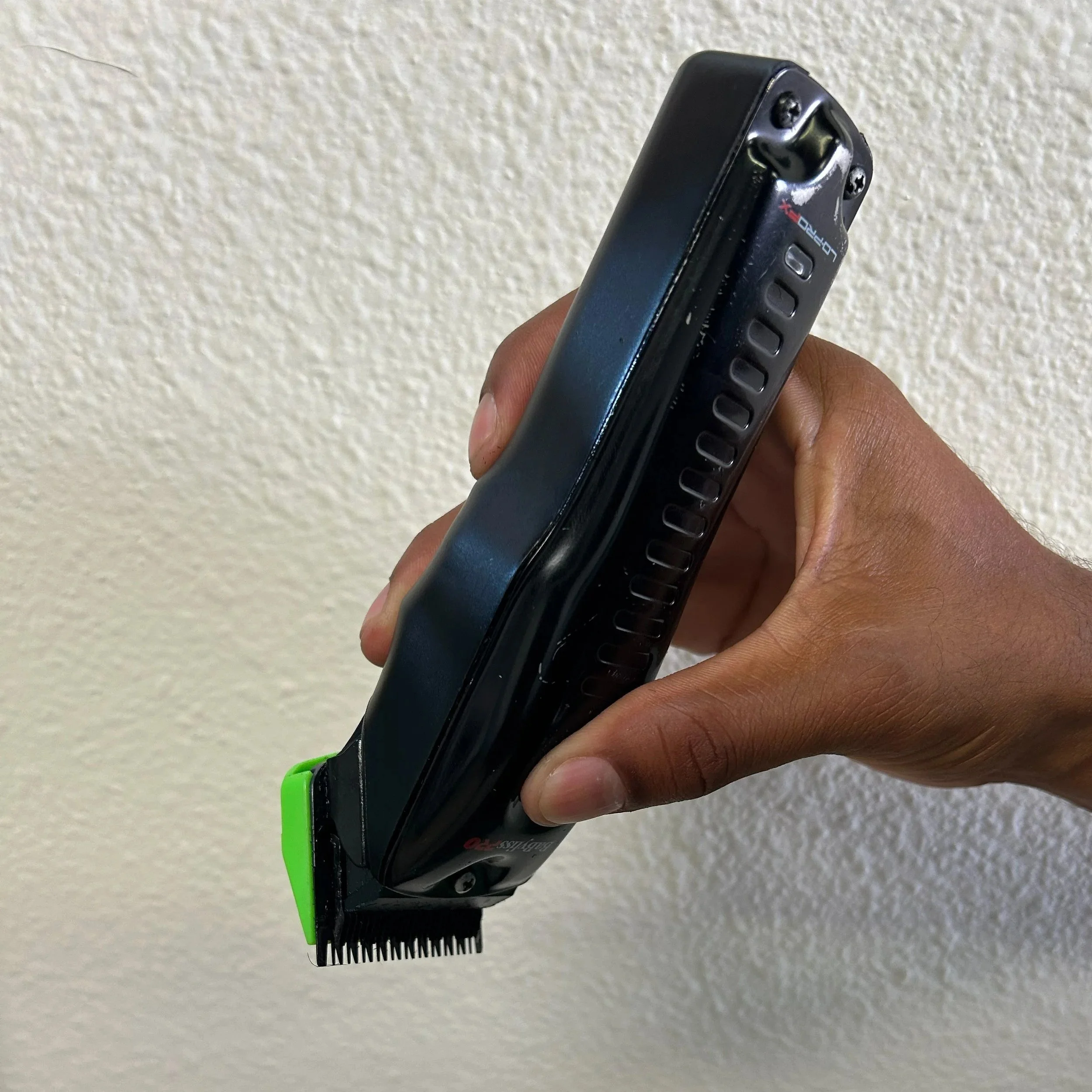 Close-up of a person's hand holding a comb and hair clippers with a green guard against a textured off-white wall.