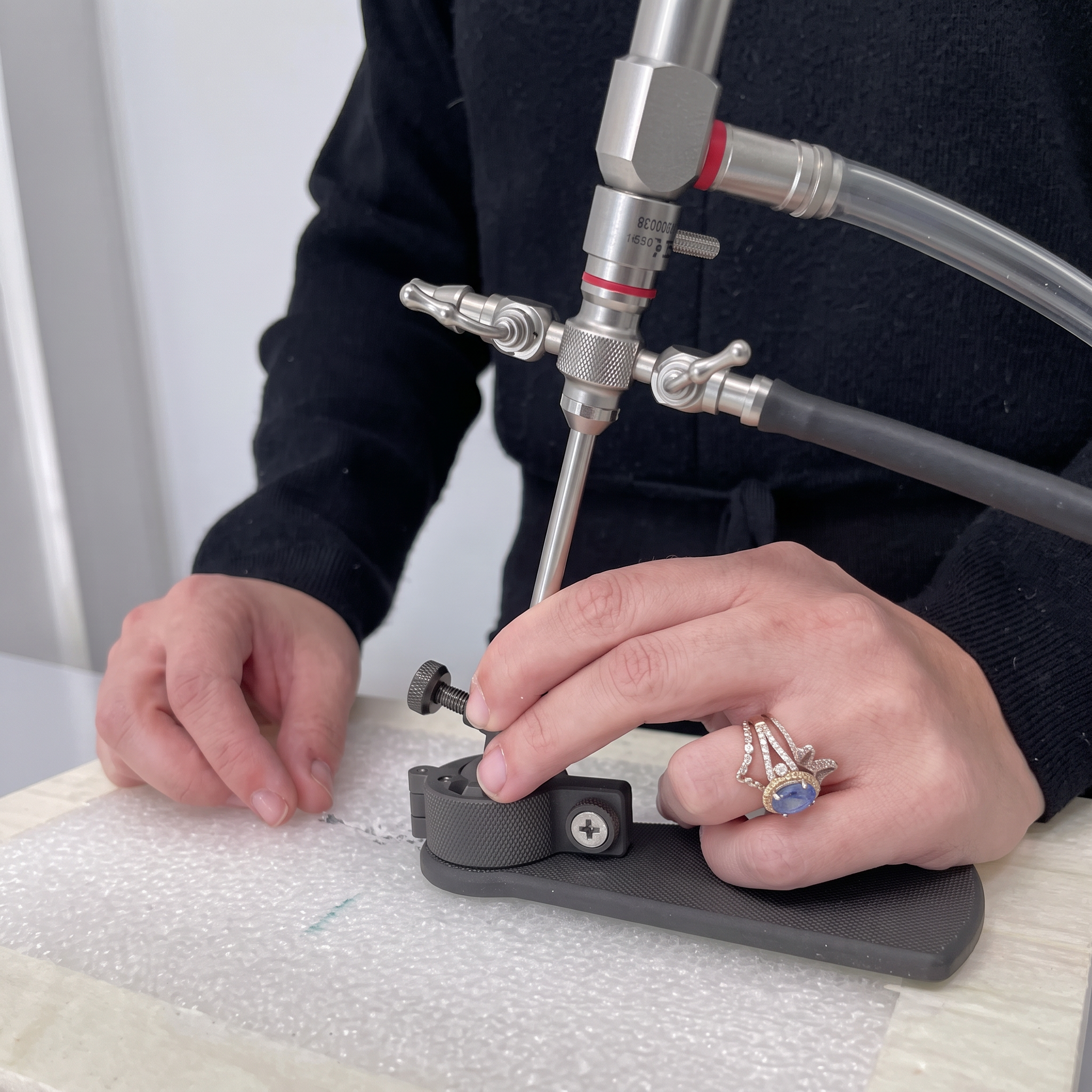 Close-up of a person's hands assembling or adjusting a metallic mechanical or medical device on a white surface, with part of the person's black clothing visible.