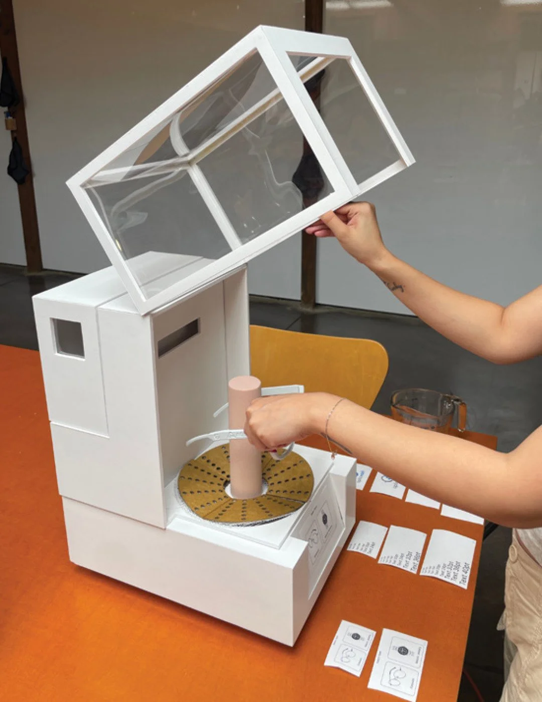 Person using a white machine with a glass lid, placing a cylindrical object inside, on an orange table.