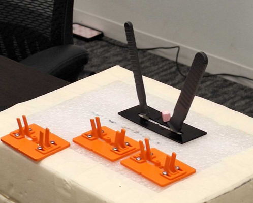 Three orange plastic router jigs with metal pins, used for woodworking, placed on a white foam board, and a separate black strip with two gray strips attached, possibly for guiding or alignment, in an indoor setting.