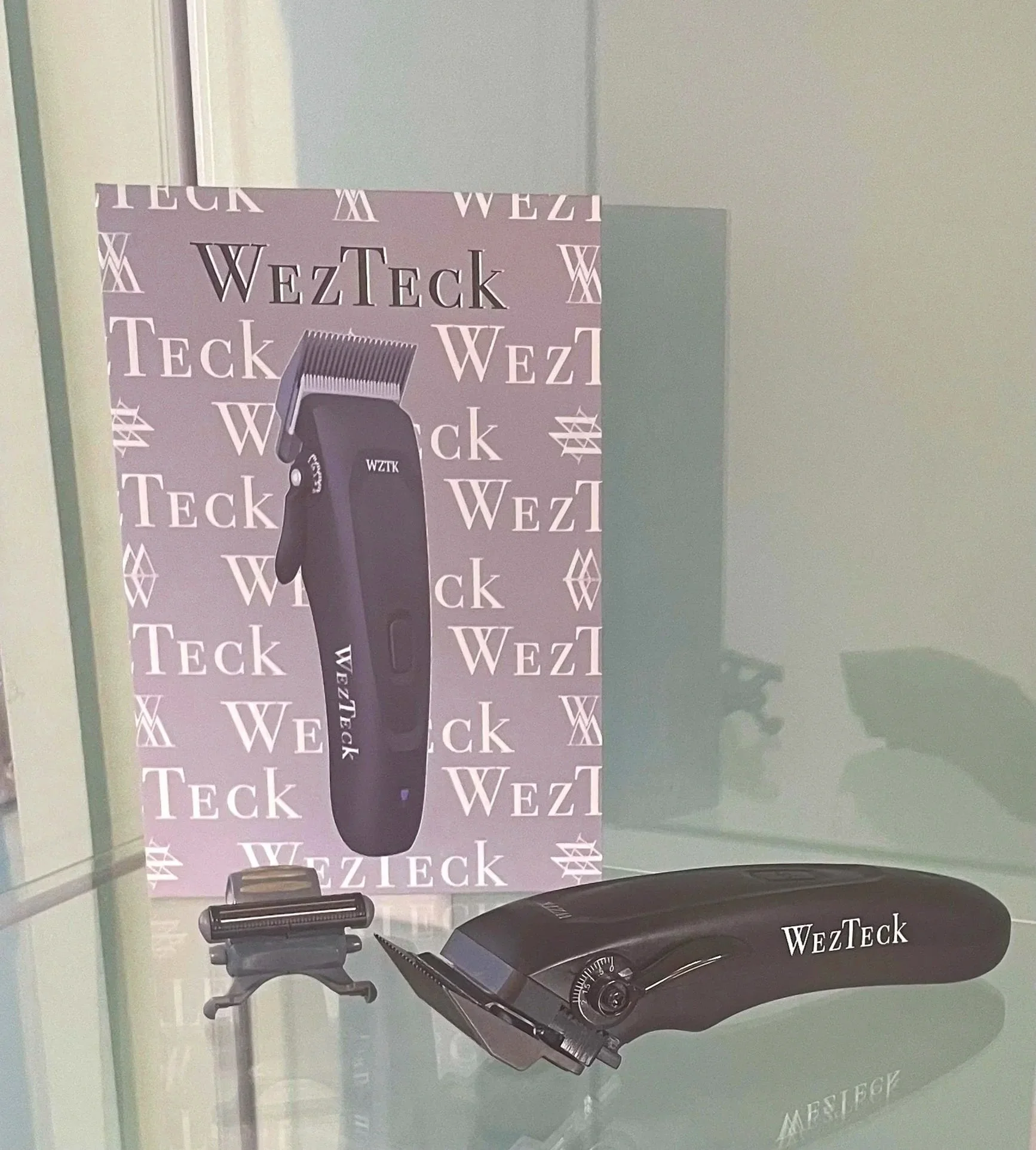 Display of a hair clipper with the brand name 'WezTeck' on a purple background and an image of a black hair clipper. The item is placed on a reflective surface, with a large WezTeck promotional poster behind it featuring the same brand name repeatedly.