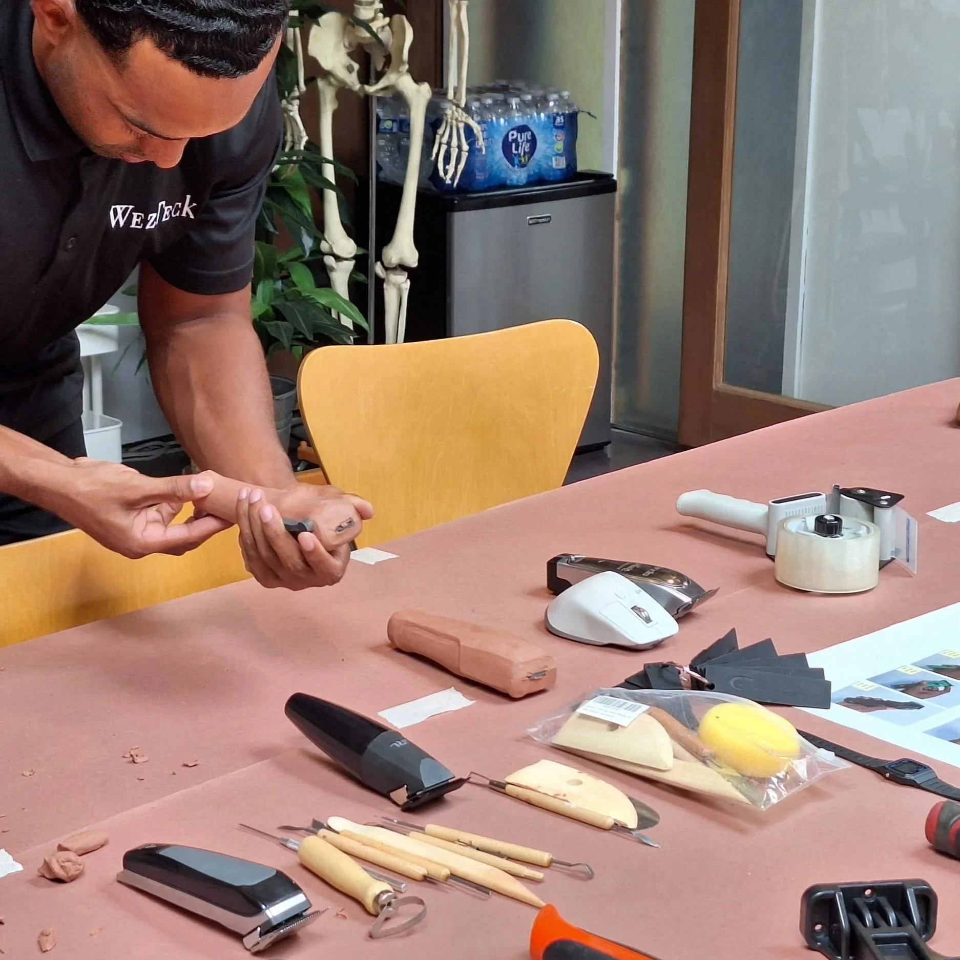 A man working on a project at a table that has sculpting tools, clay, and other art supplies. In the background, a skeleton model and a pack of bottled water are visible.