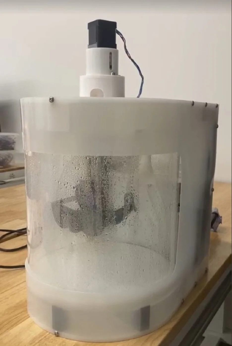 A laboratory machine with a transparent front panel showing condensation and a mechanical arm inside, placed on a wooden table.