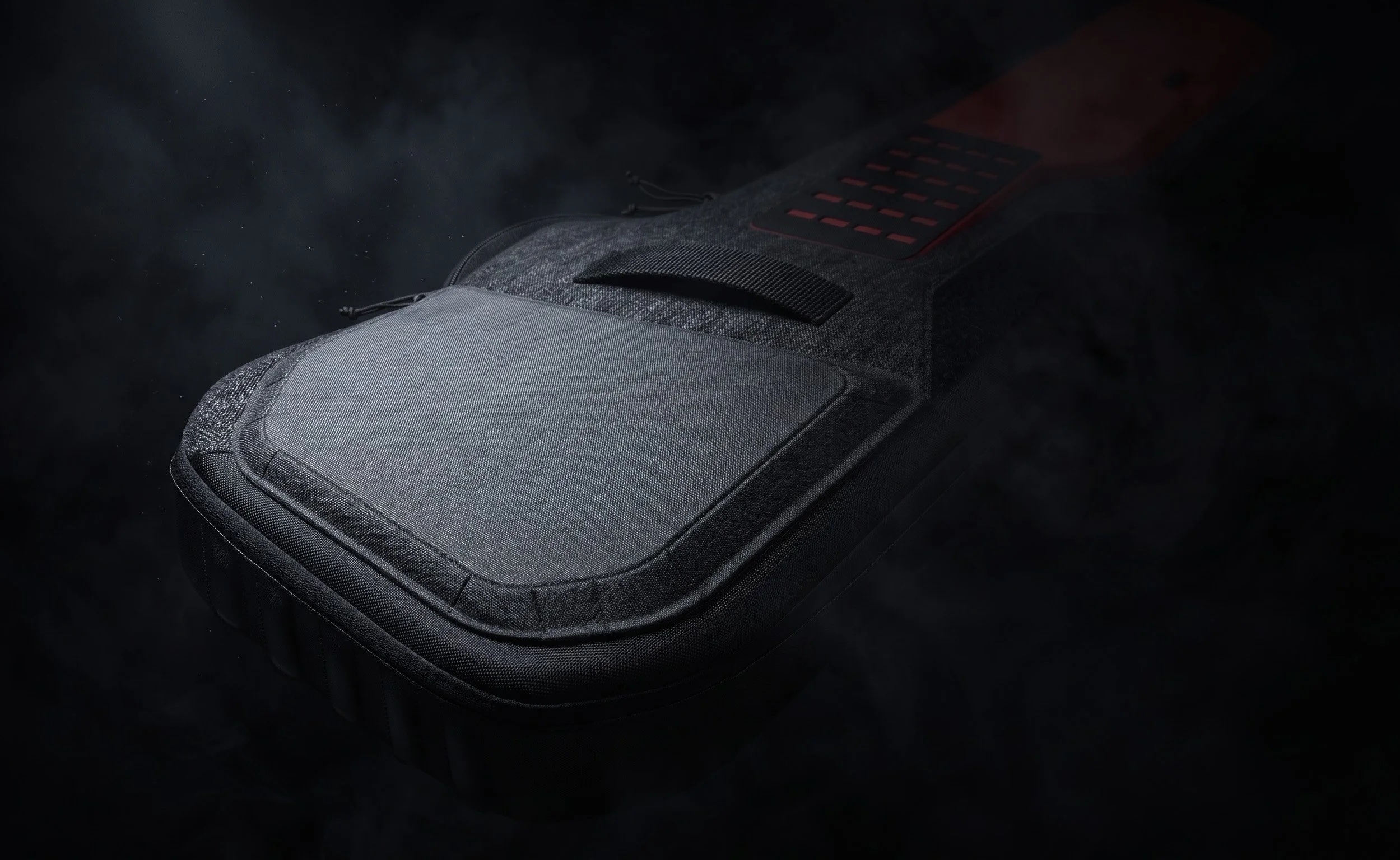 A dark-colored backpack with a textured fabric, black handle, and red accents, isolated on a black background with dust particles visible in the air.