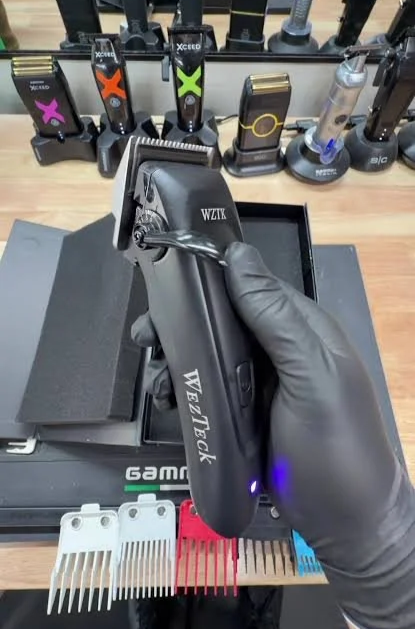 A person wearing black gloves holding a black wizard haircutting clipper over a black grooming station. In the background, there are various clipper blades and attachments arranged on a shelf.