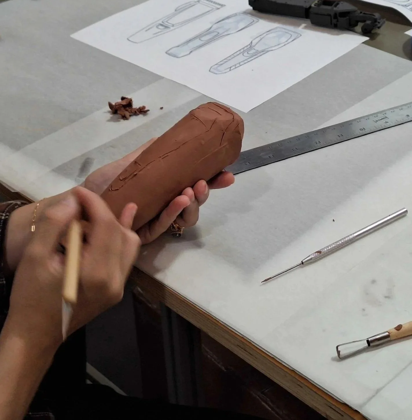 Person holding a rectangular piece of clay on a work table, designing or shaping it with a tool; the table has drawing sketches of objects, a ruler, many drafting tools, and paper.