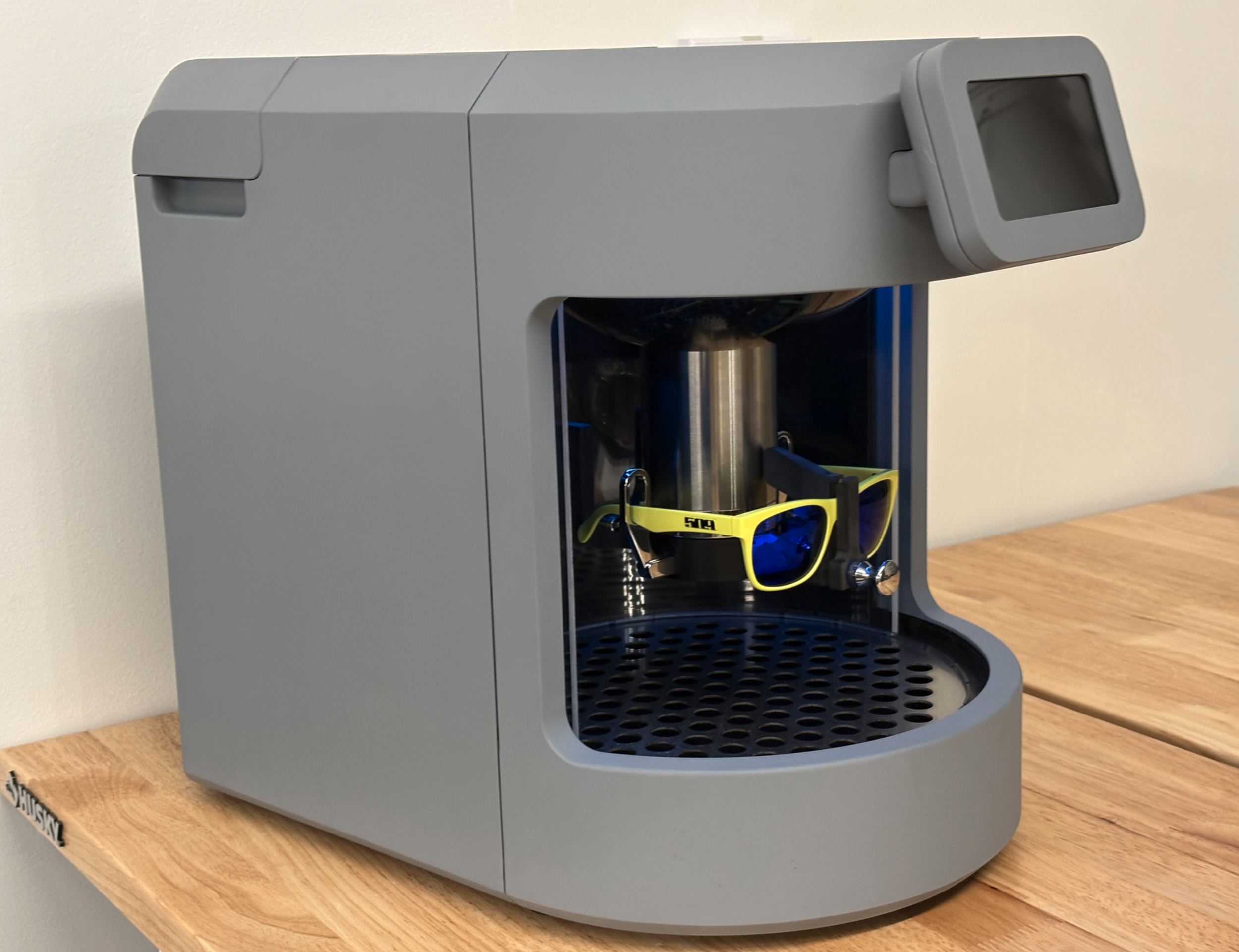 A gray 3D printer with an open front showing a cylindrical metal object being printed. There are safety glasses placed on the printer