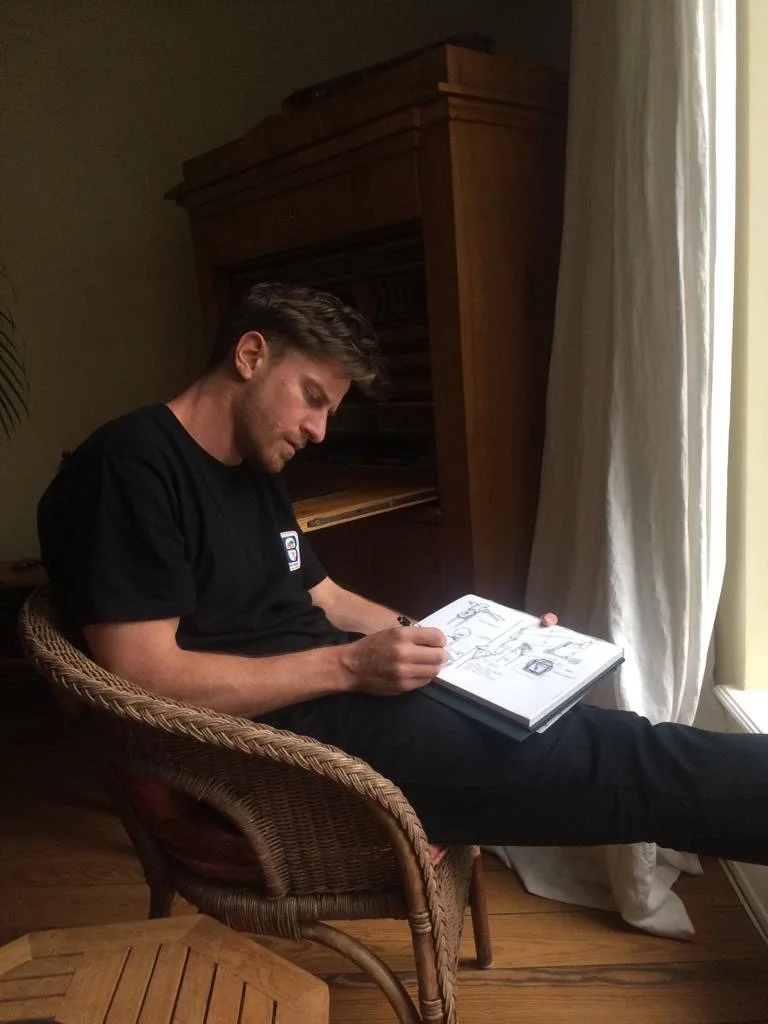 A young man with brown hair sitting in a wicker chair, drawing in a sketchbook. He is wearing a black t-shirt and black pants. The setting appears to be indoors, near a window with white curtains, and there is a wooden cabinet behind him.