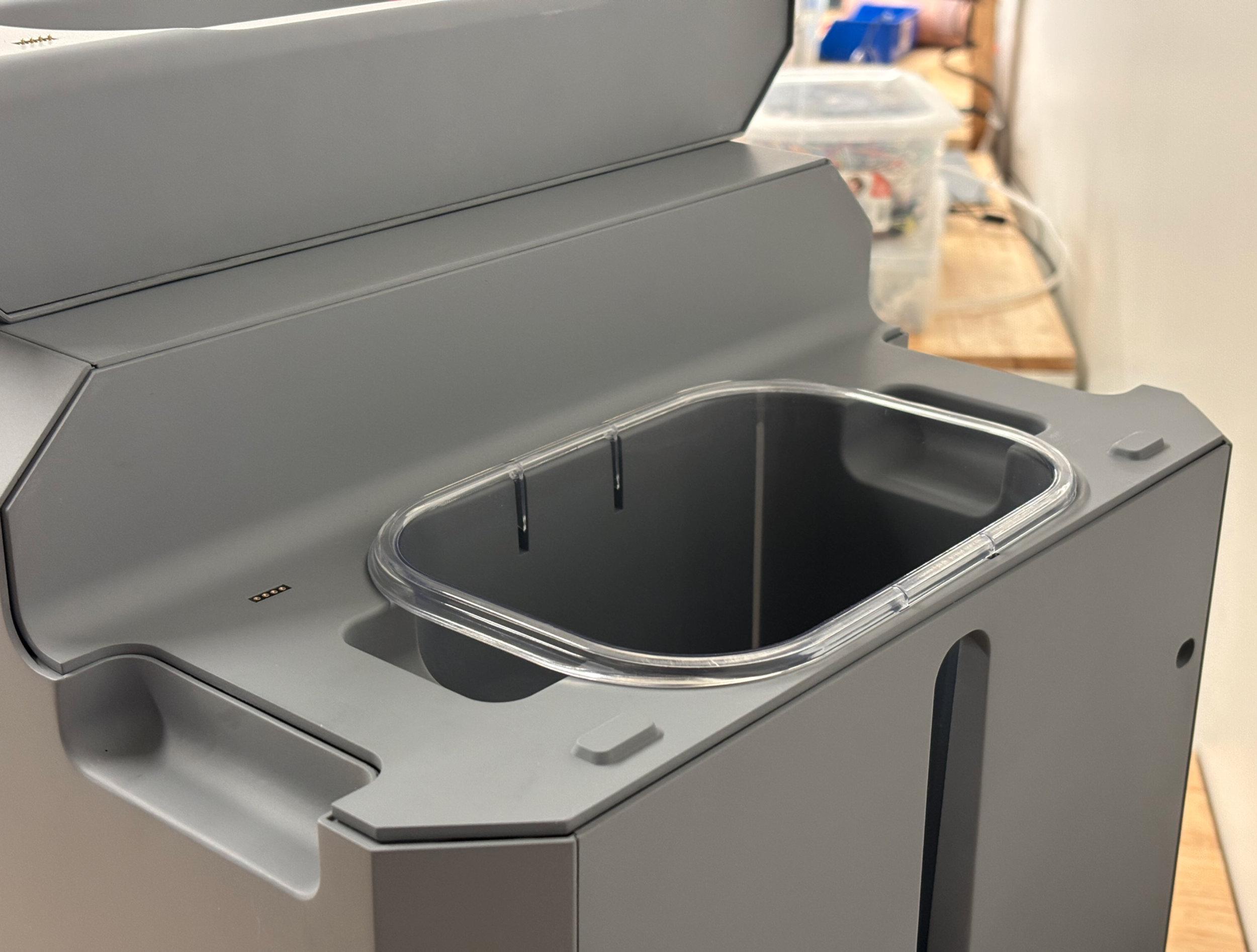 A gray laboratory or medical device with an oval opening and a clear plastic cover, set on a surface with other equipment visible in the background.