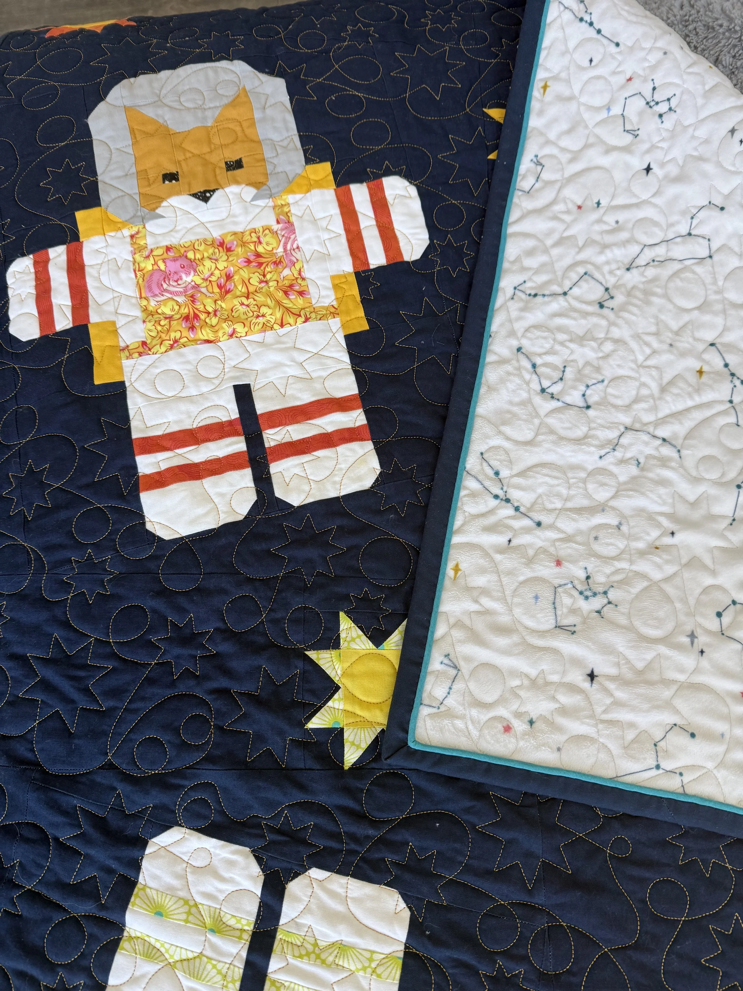 Close-up of a quilt with kitty astronauts. The quilt features a central design of an astronaut in a suit made of various colorful fabrics, set against a dark blue background with quilted star and circle patterns.