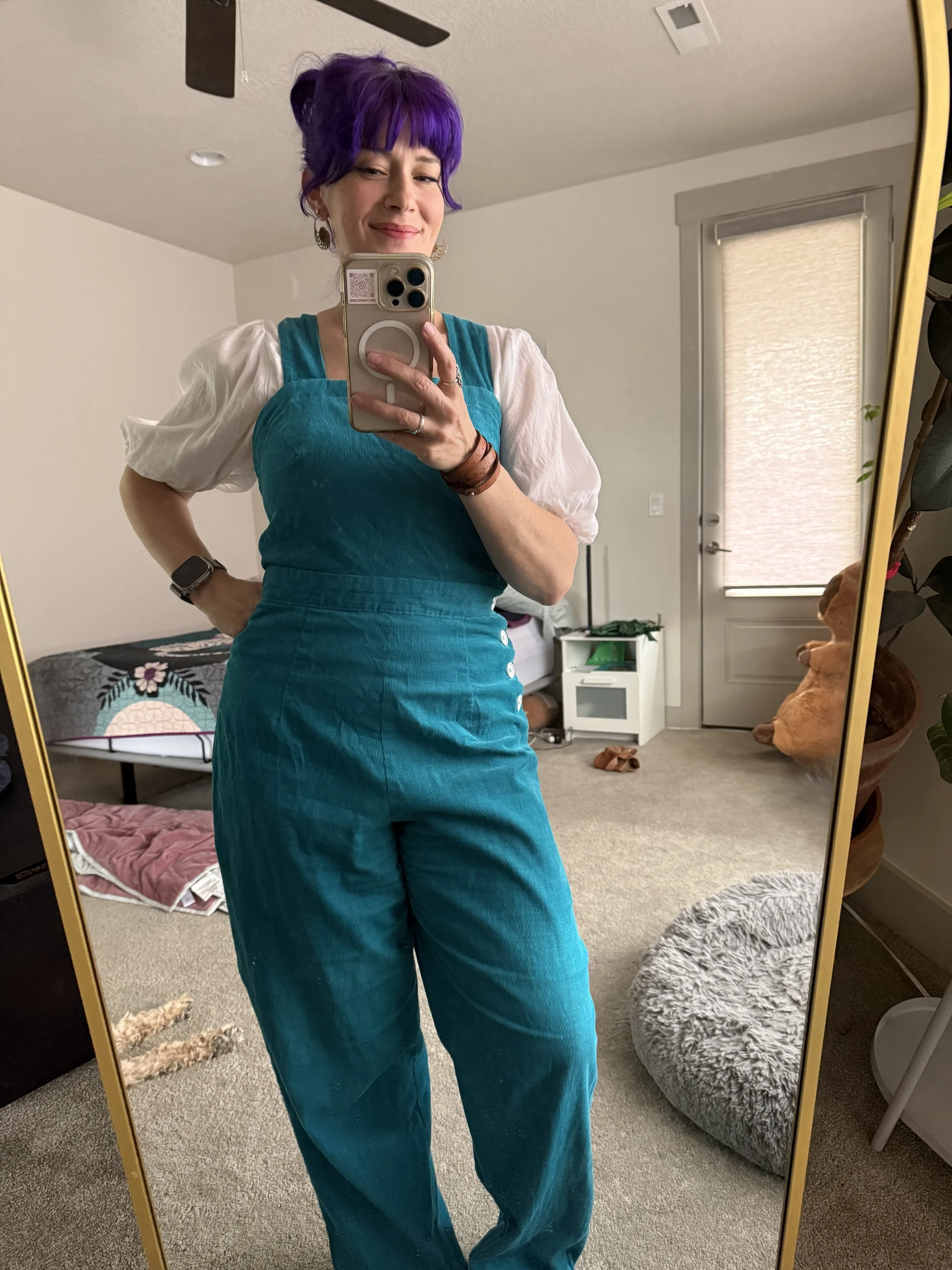 A woman with purple hair taking a mirror selfie in a bedroom, wearing a teal jumpsuit with a white puffy-sleeve shirt underneath, a black wristwatch, and earrings.