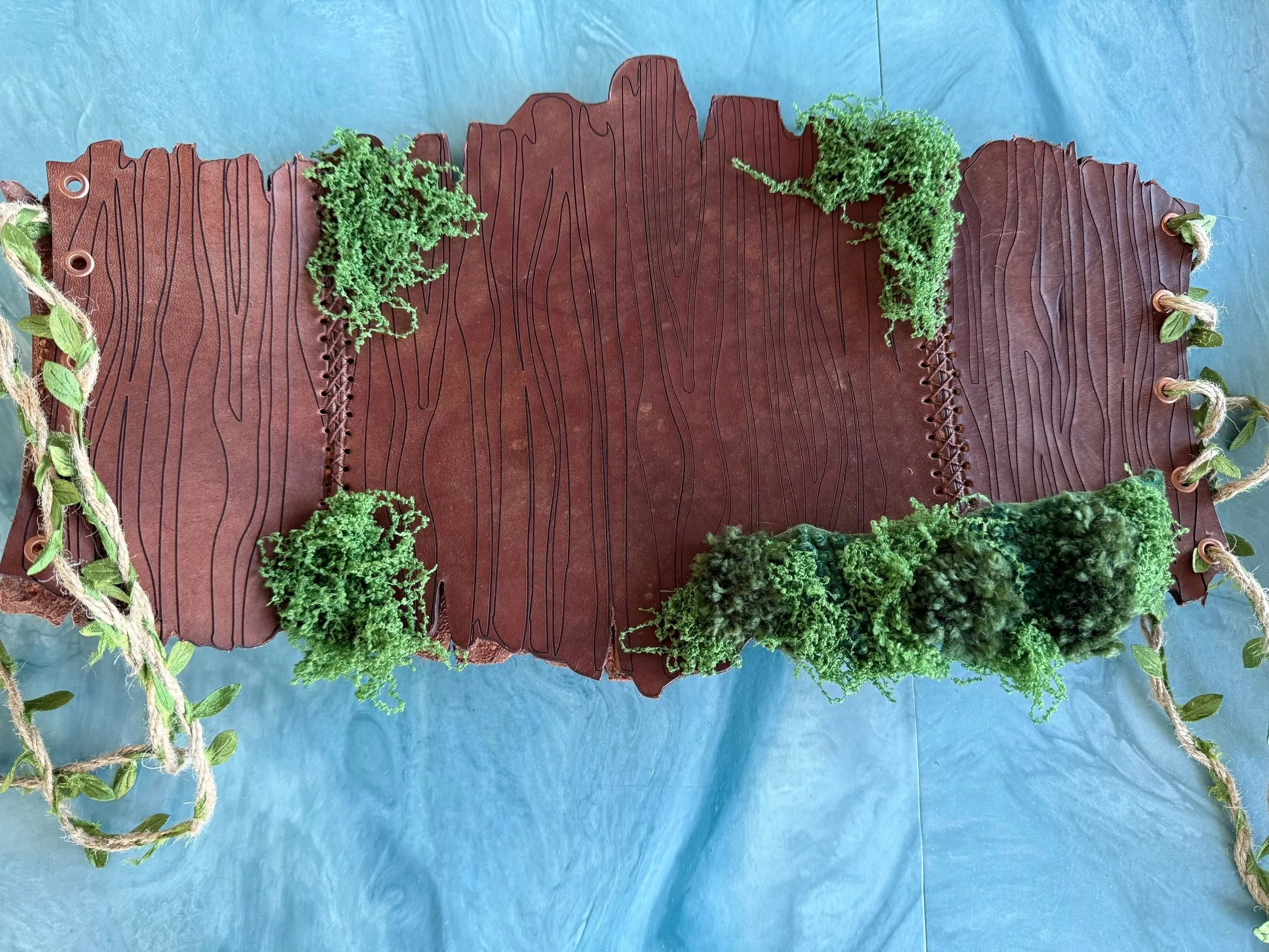A leather corset belt featuring a faux wooden design, decorated with embroidered moss, side ties are a jute rope with green leaves woven through it.