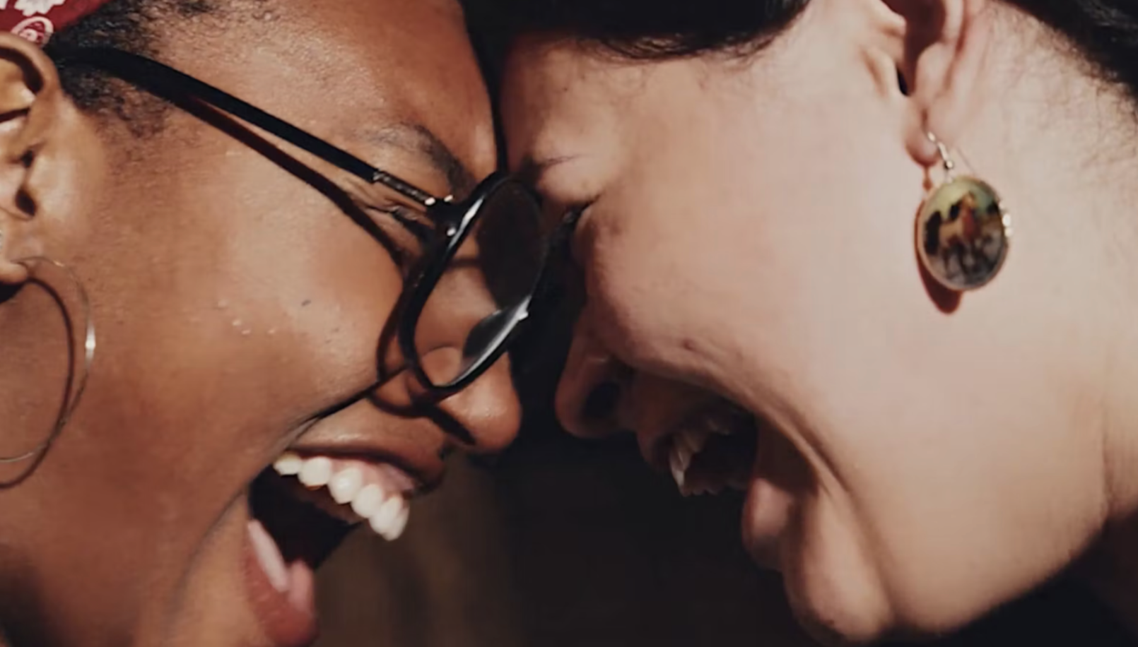 Two women are close together, smiling and laughing with their foreheads touching. One is wearing glasses and hoop earrings, while the other has earrings with a rainbow-colored design. Their faces show joy and connection.