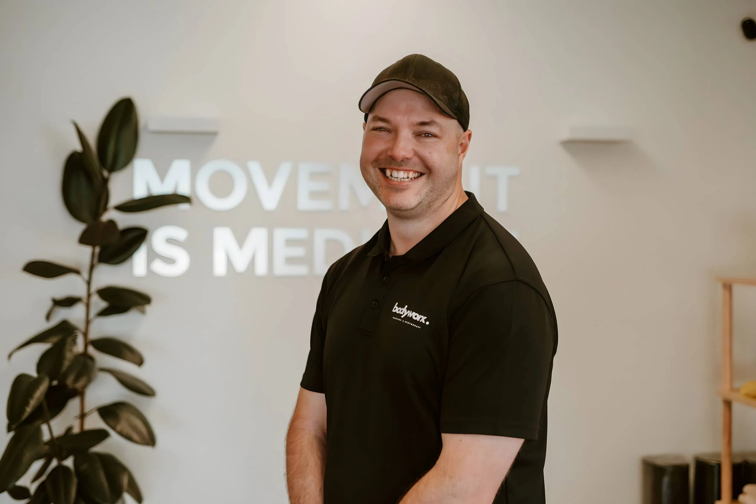 A smiling man wearing a black polo shirt and a brown cap stands indoors. Behind him, there is a large plant on the left side and a wall with a blurred sign that reads 'MOVEMENT IS MEDIC...'.
