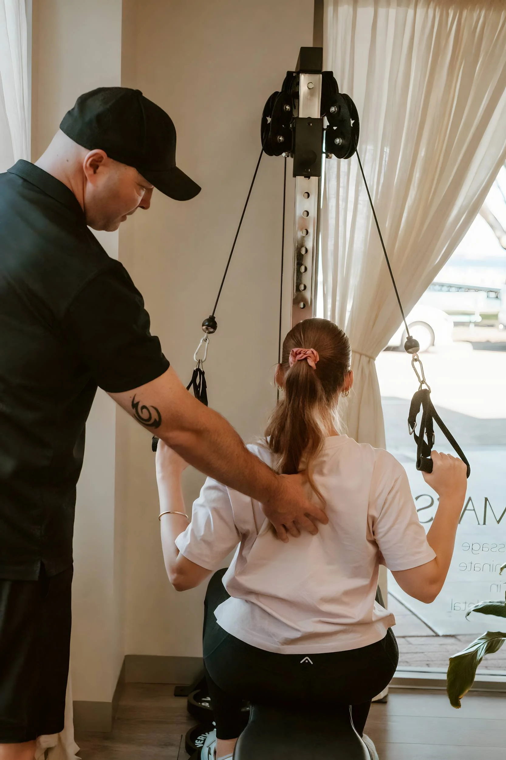 A woman sitting on a black chair on a wooden floor, receiving physical therapy or chiropractic care from a man. The man is standing, wearing a black cap and black shirt, assisting her with a neck support device attached to a pulley system near a window with curtains.