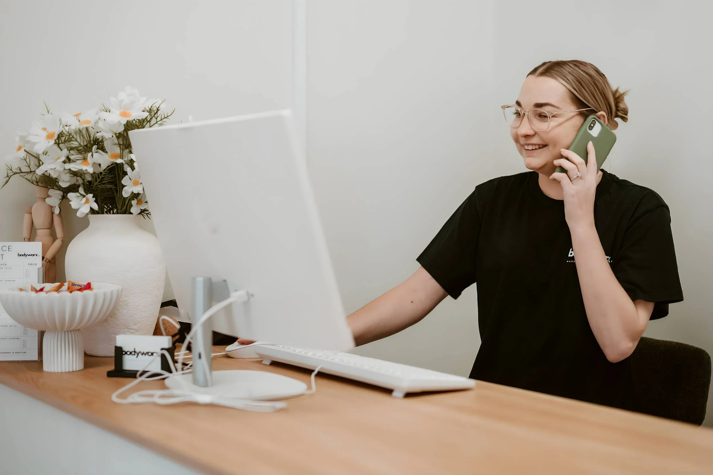 A woman with glasses smiling and talking on the phone at her desk, which has a white computer monitor, keyboard, and mouse, with vase of white flowers and a bowl of candy.