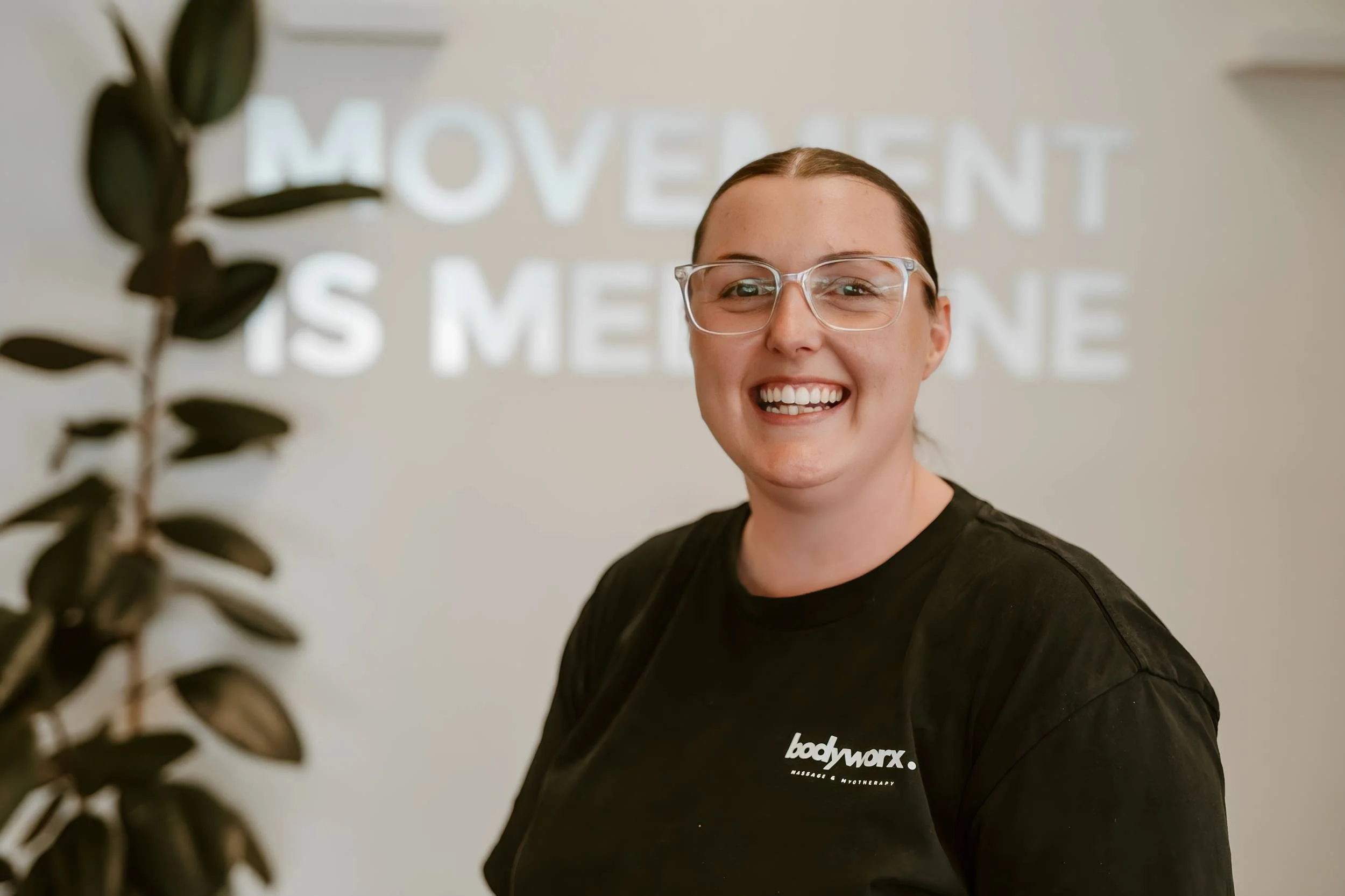 Smiling woman with glasses and dark hair in a black shirt with 'bodyworx' logo stands in front of a wall with the word 'MOVEMENT' and other text blurred in the background.
