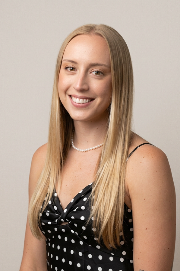A young woman with long blonde hair, fair skin, wearing a black dress with white polka dots, a pearl necklace, and smiling at the camera against a plain light gray background.