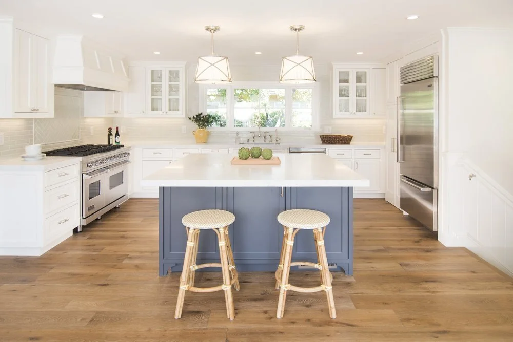 Bright, modern kitchen with white cabinets, a blue island, wooden flooring, and stainless steel appliances, including a refrigerator and stove. Two pendant lights hang above the island, which has a wooden tray with green decorative balls and two wicker bar stools.