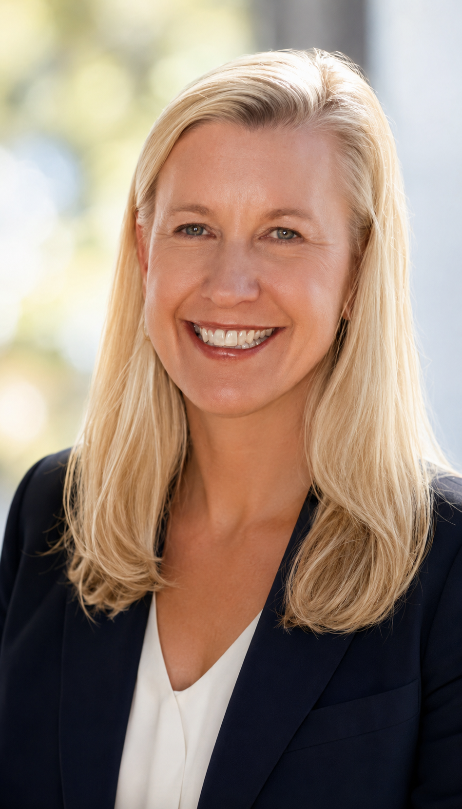 A woman with blonde hair smiling, wearing a dark blazer and white blouse, in front of a blurred outdoor background.