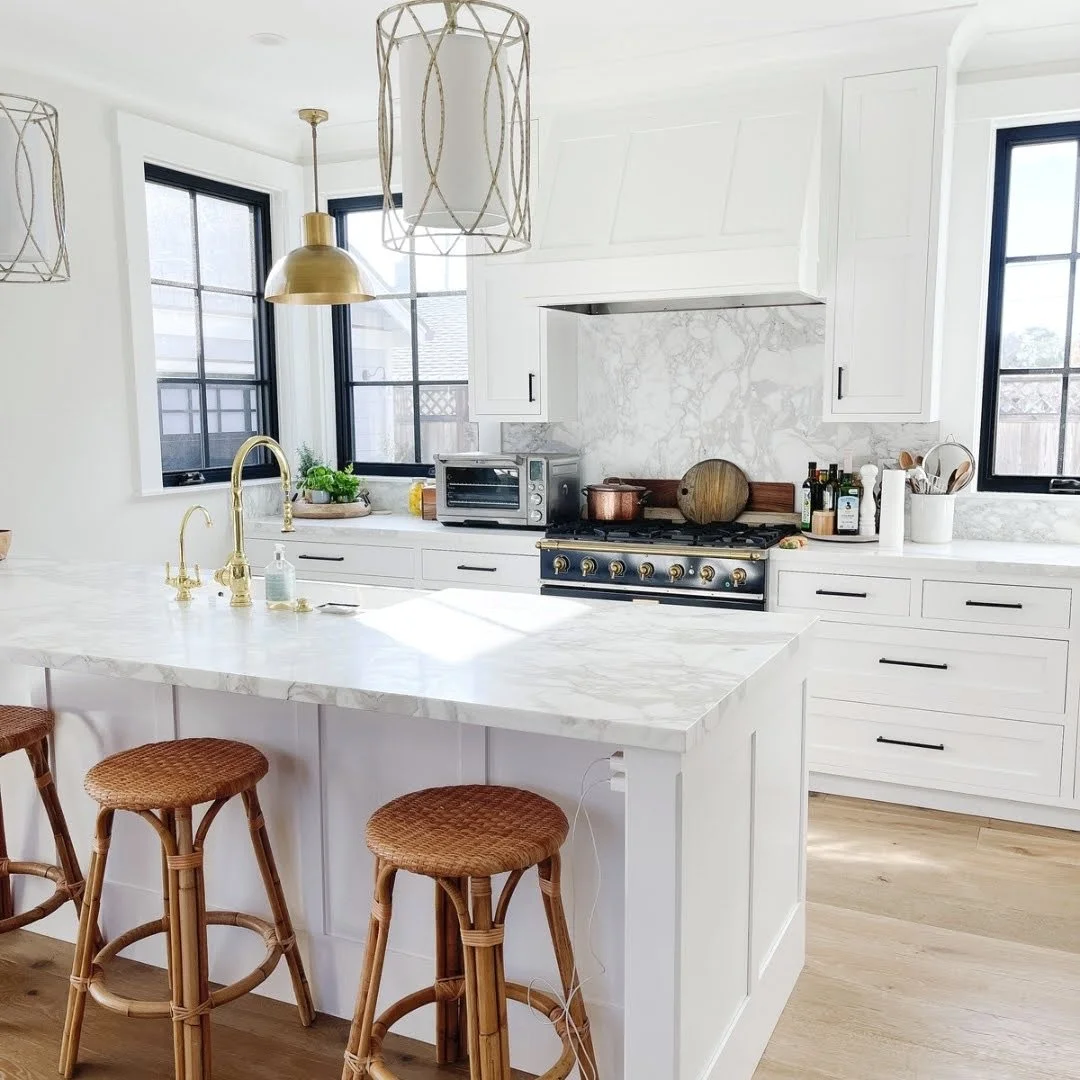 Modern white kitchen with marble island, gold fixtures, black window frames, and wicker stools.