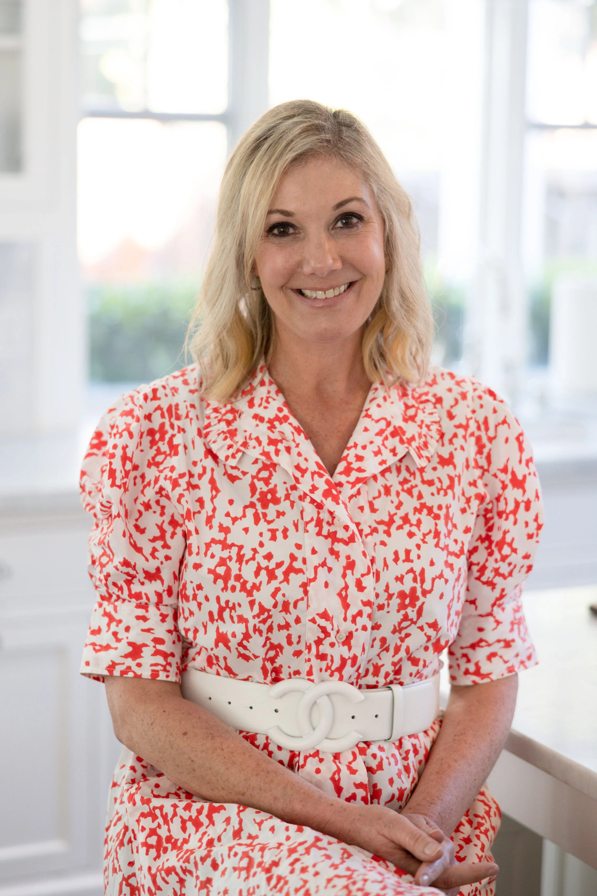 A woman with blonde hair wearing a red and white patterned dress smiling, sitting by a window with natural light.