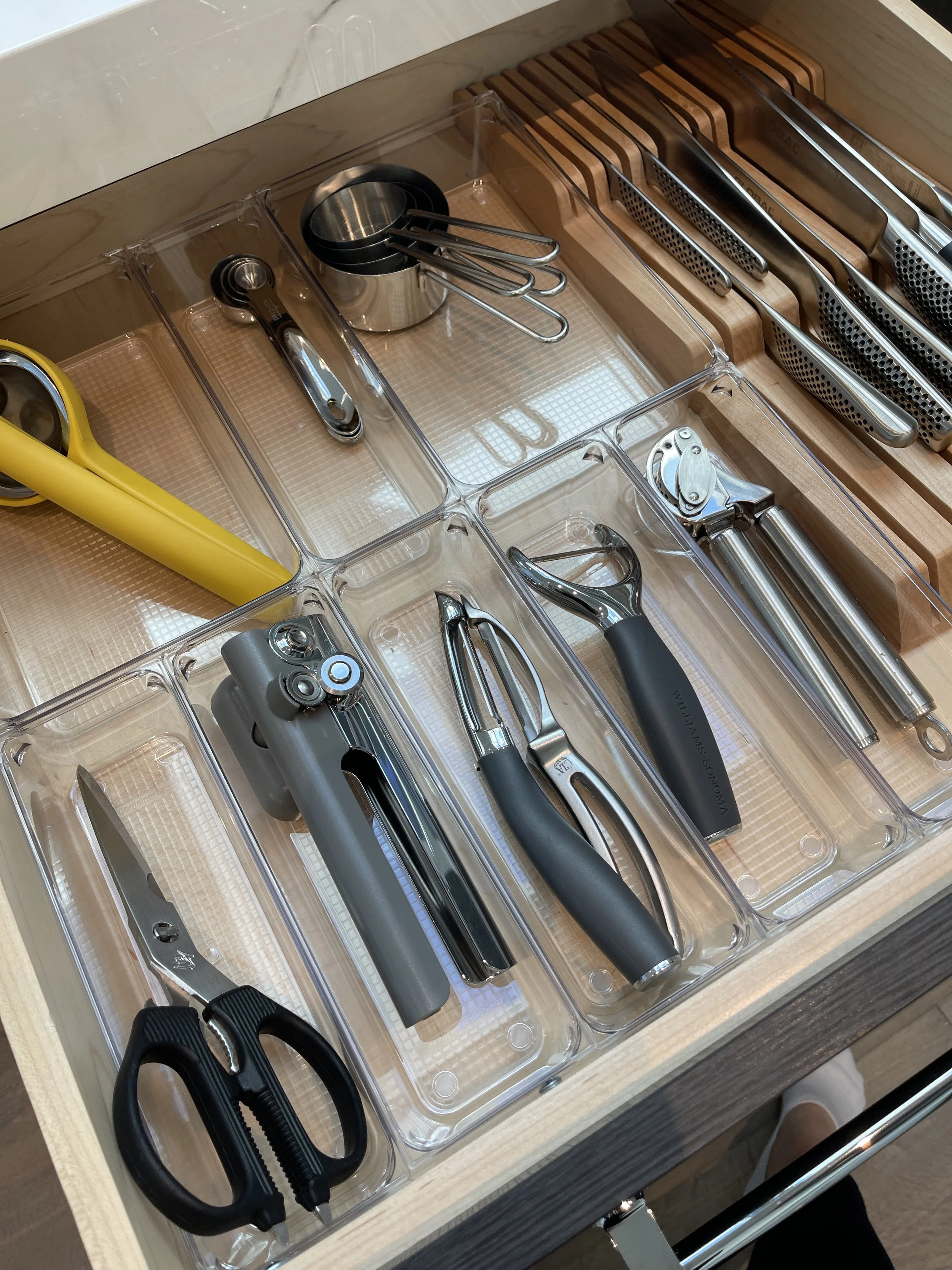Kitchen drawer organized with kitchen tools including scissors, a measuring cup, garlic press, can opener, and knives in wooden and metal handles, all stored in clear plastic compartments.