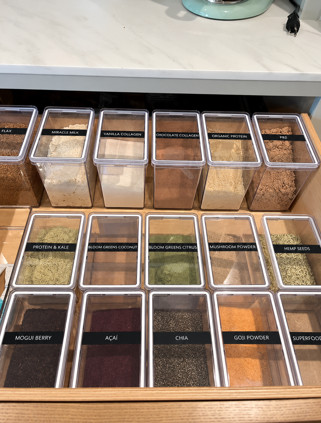 A collection of labeled health food powders in clear containers, including flax, miracle milk, vanilla collagen, chocolate collagen, organic protein, PB2, protein & kale, bloom greens coconut, bloom greens citrus, mushroom powder, hemp seeds, mogui berry, acai, chia, goji powder, and superfood.