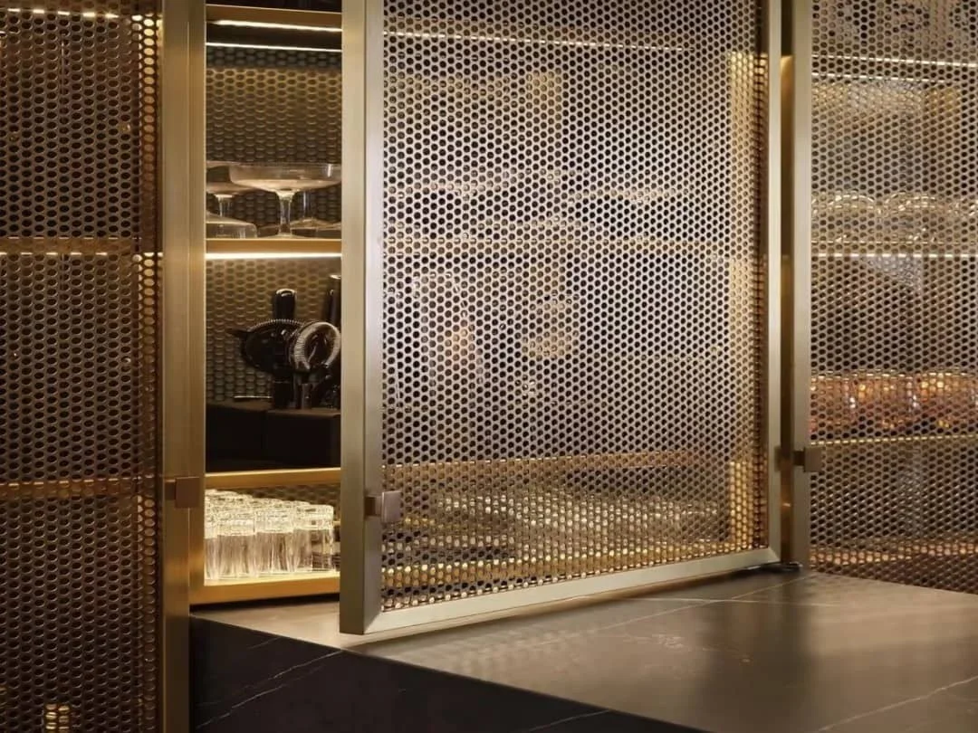 A gold-colored perforated metal cabinet with sliding doors, containing glassware and a coffee machine visible through one door.