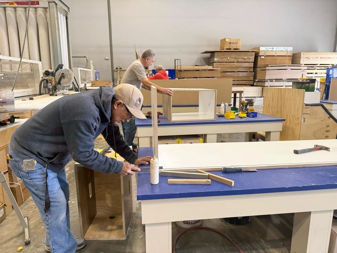 Three men working on woodworking projects in a large workshop with various tools and stacks of wood in the background.