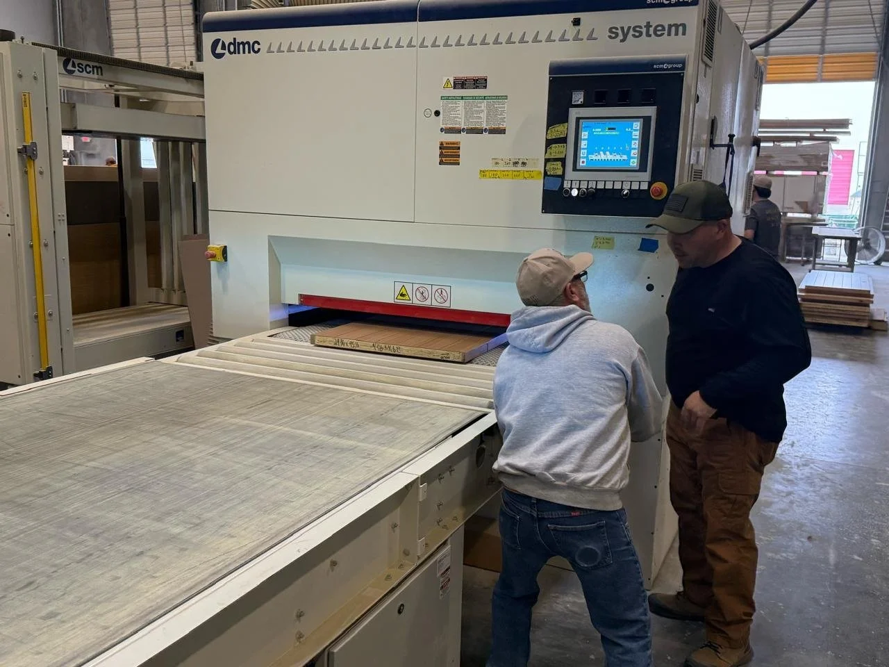 Two men talking near a large industrial woodworking machine in a factory, with wood panels and stacks of wood in the background.