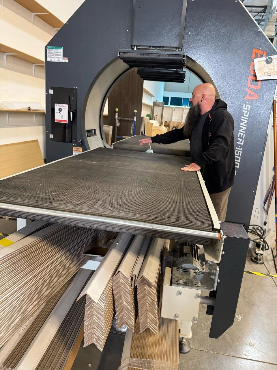A man with a beard operating a large industrial CNC machine in a woodworking shop, with shelves and wood pieces in the background.