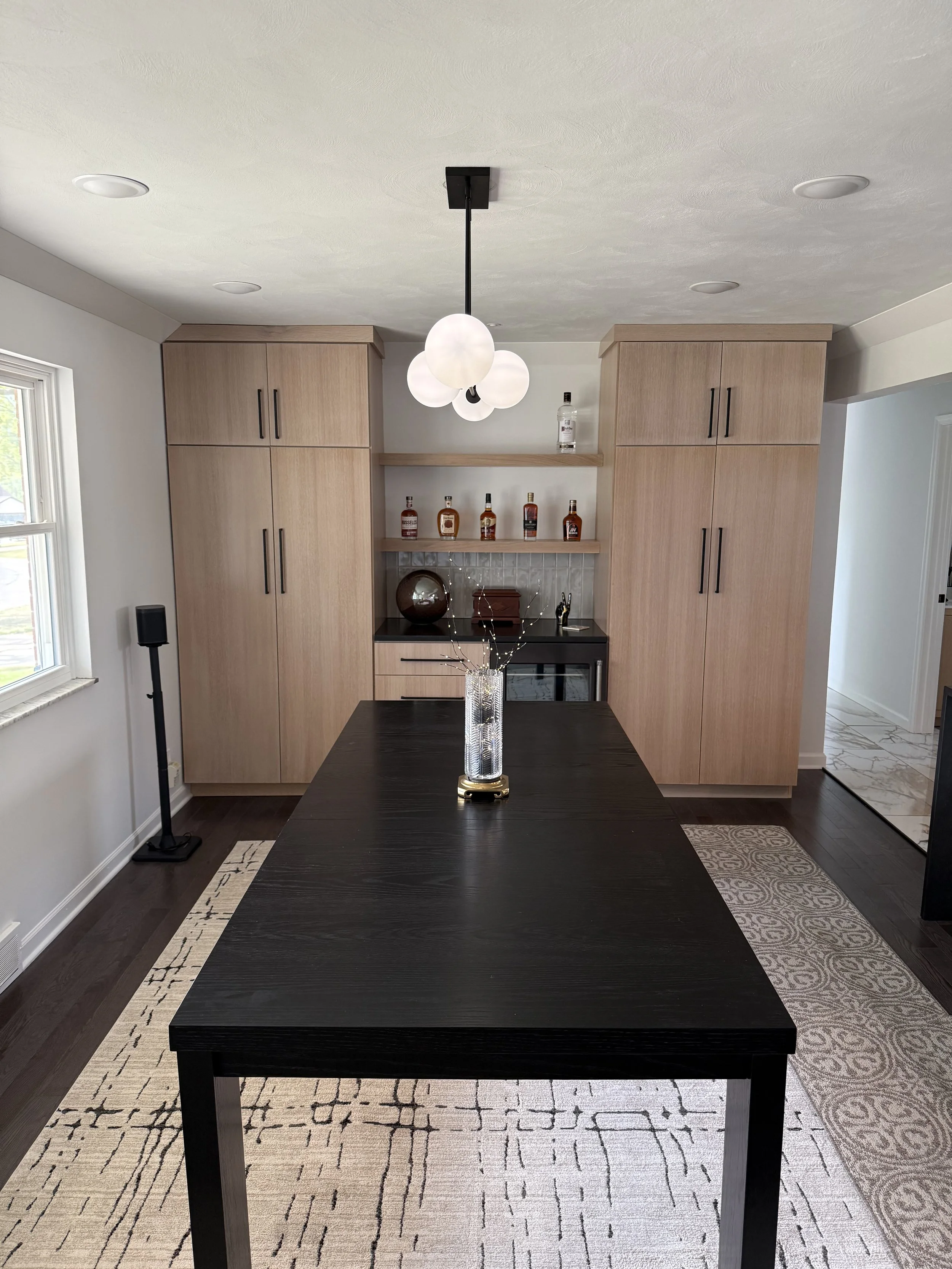 A dark wooden dining table in a room with built-in wooden cabinets. A modern pendant light with multiple white globes hangs above the table. The room has a window on the left side and an area rug underneath the table.