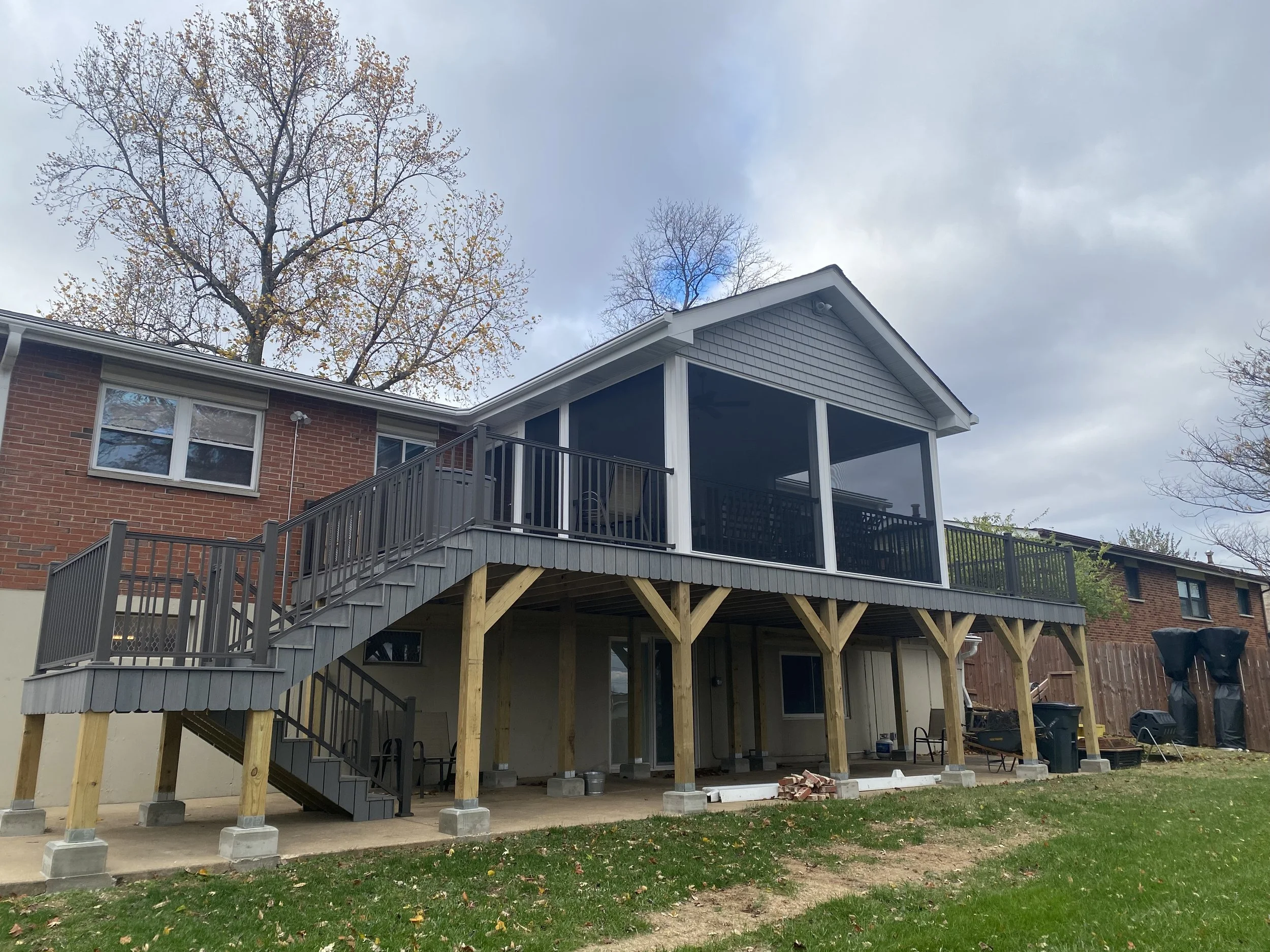 Back of a two-story brick house with a new elevated deck under construction, with stairs, railing, and support beams, surrounded by trees and a cloudy sky.