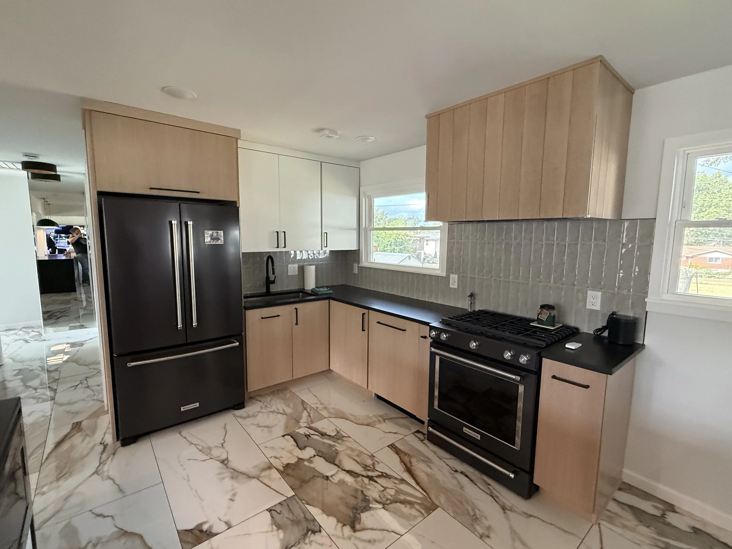 Modern kitchen with black refrigerator, black stove, wooden cabinets, marble tile floor, and two windows letting in natural light.