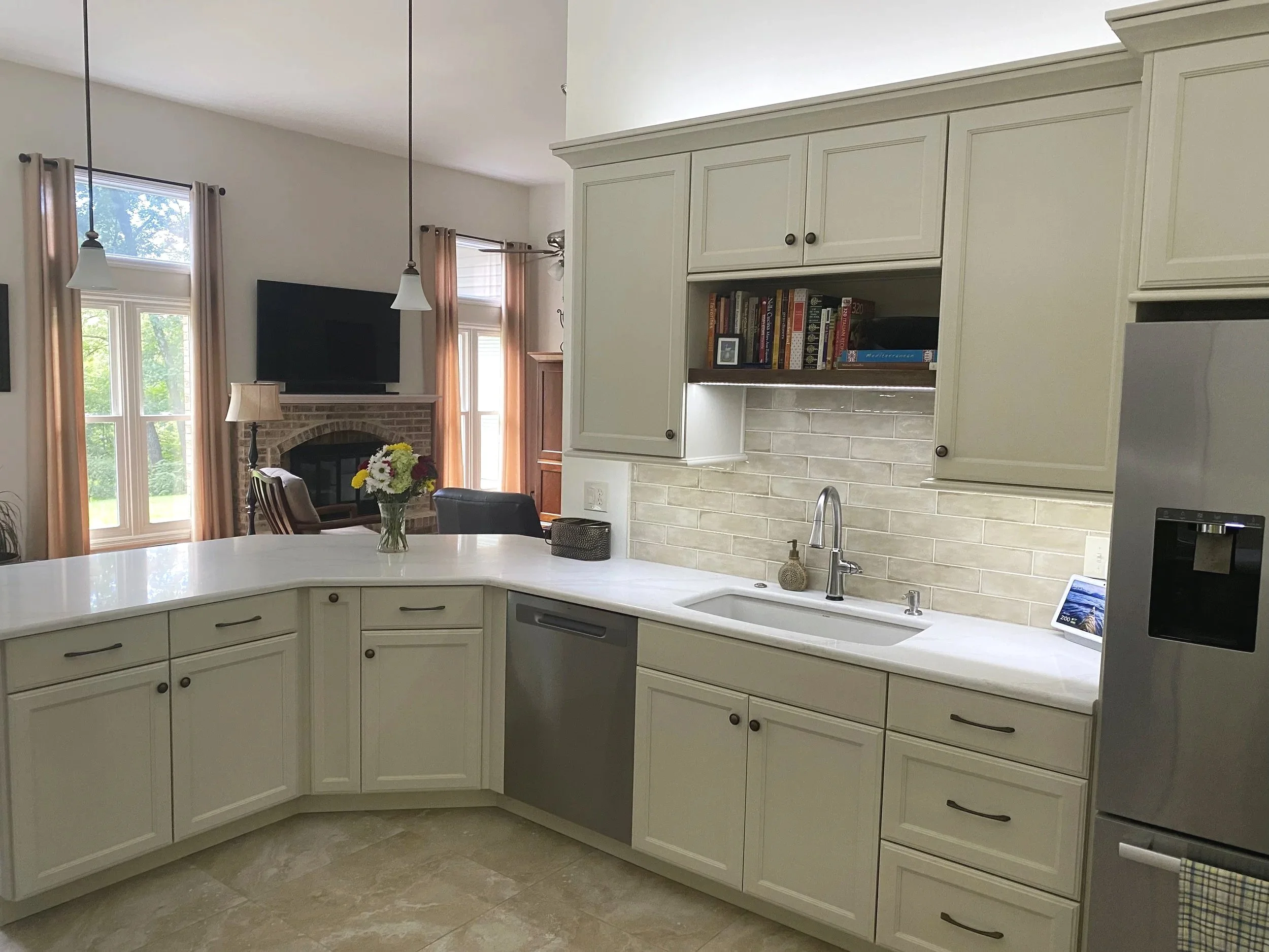 Open kitchen with white cabinets, a white countertop, a sink with a faucet, and a decorated island. In the background, there is a living room with a fireplace, a TV, large windows with beige curtains, and a vase with flowers on the counter.