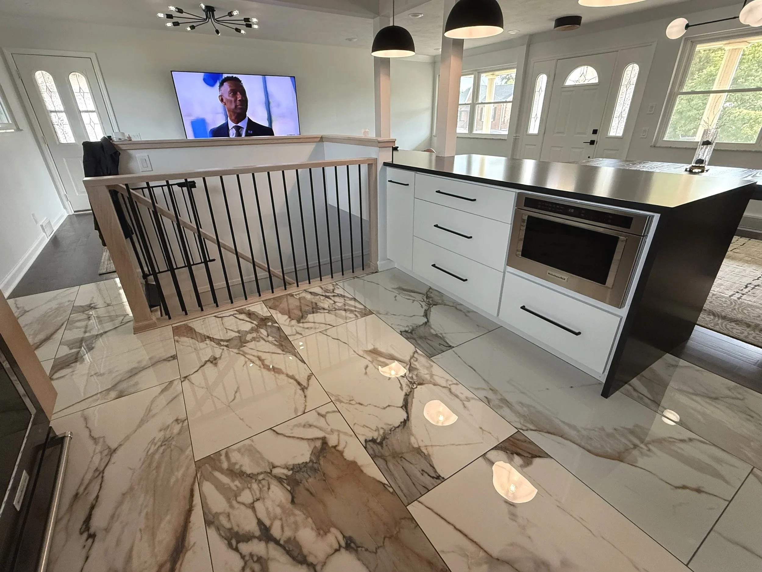 Interior view of a modern kitchen with marble floor tiles, white cabinets, a black countertop, and a microwave oven. There are black and white hanging pendant lights and a large window letting in natural light. A television is mounted on the wall, and part of a staircase with black railings is visible.