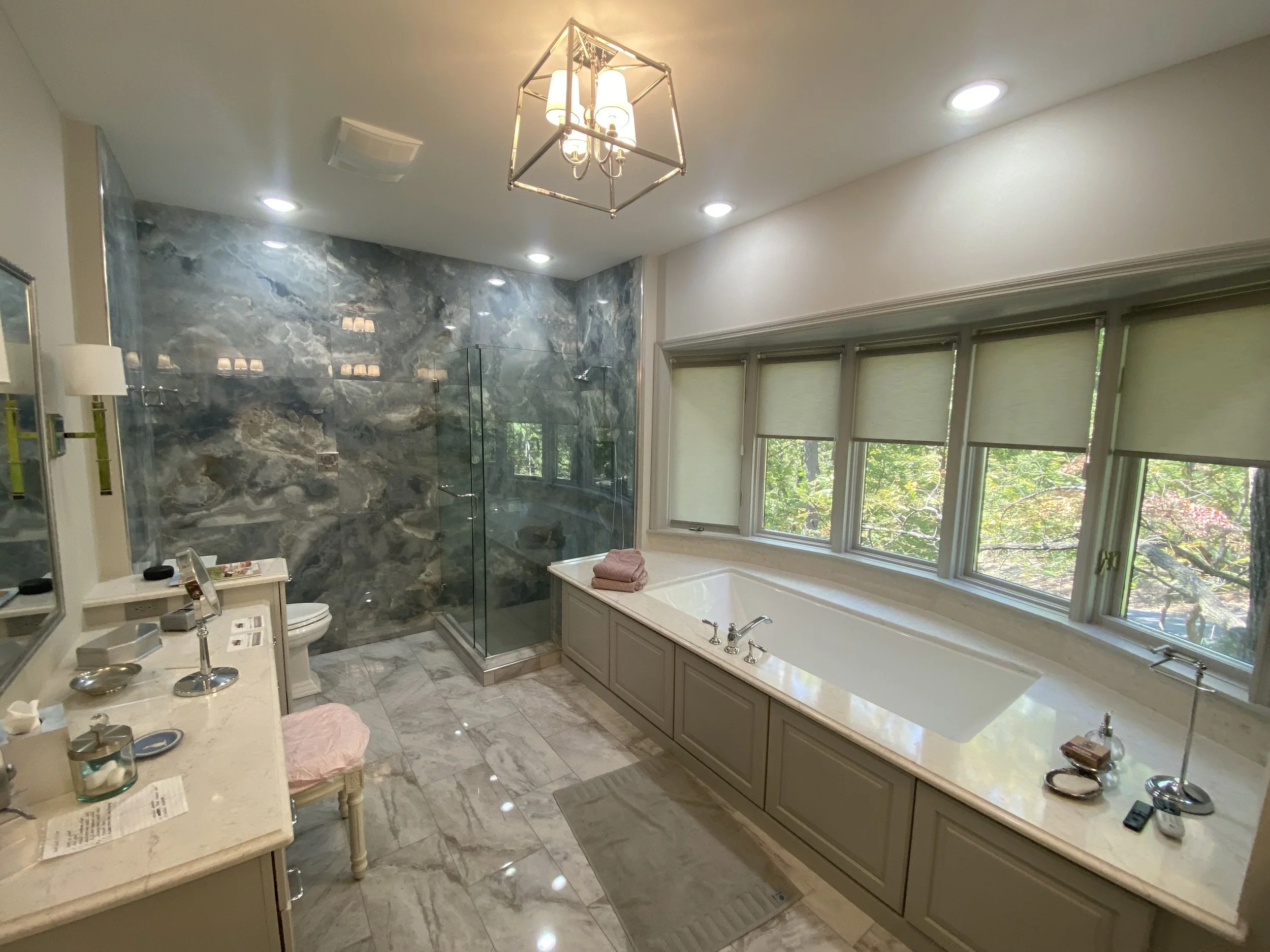 A spacious bathroom with a large window showing trees outside. It features a marble shower with a glass door, a bathtub beneath the window, and multiple white and beige fixtures. There is a chandelier hanging from the ceiling and recessed lighting.