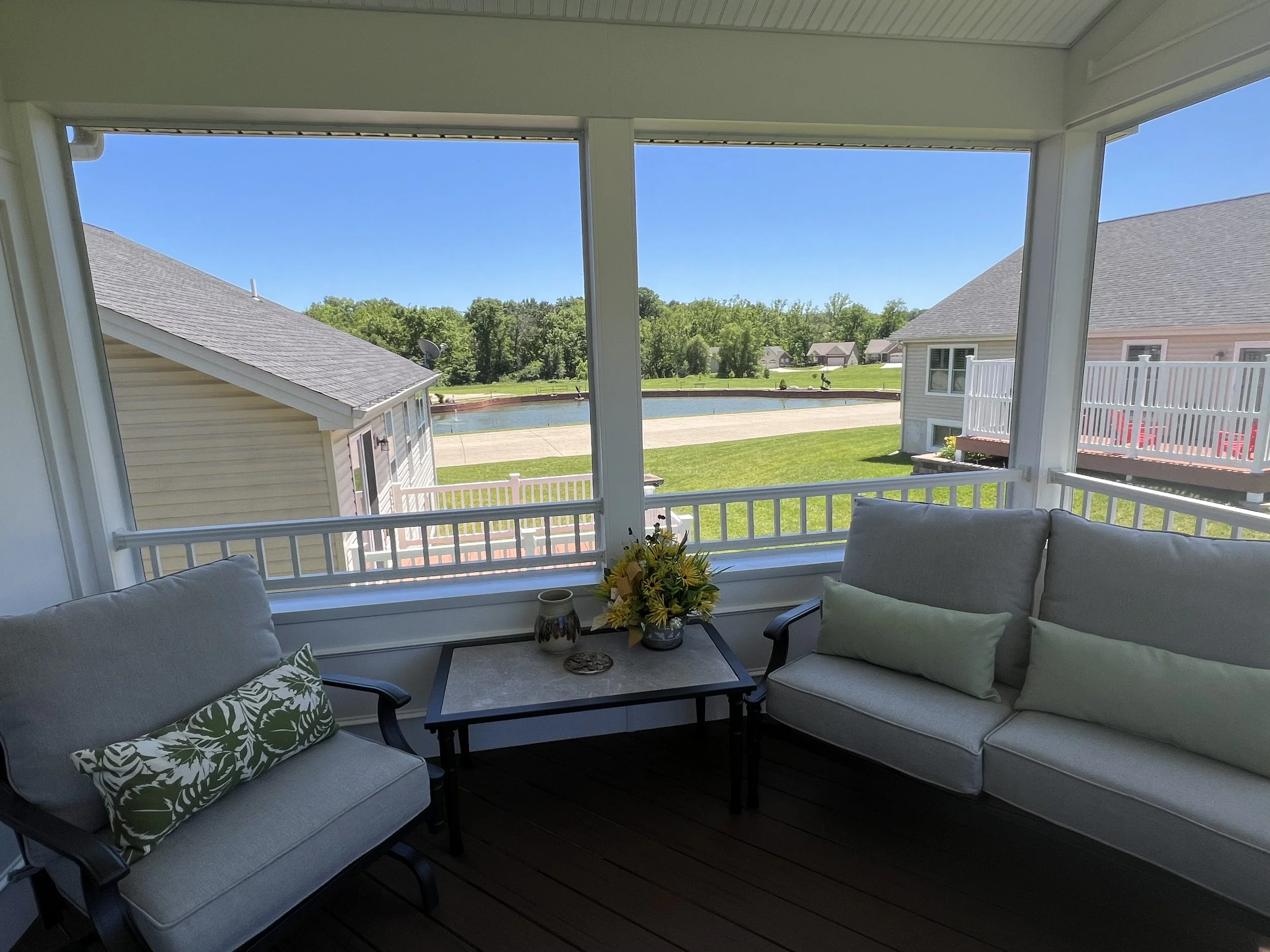 View from a screened porch with beige cushioned furniture, a small side table with a flower arrangement, and a scenic view of a backyard with a pond, green lawn, and houses under a clear blue sky.
