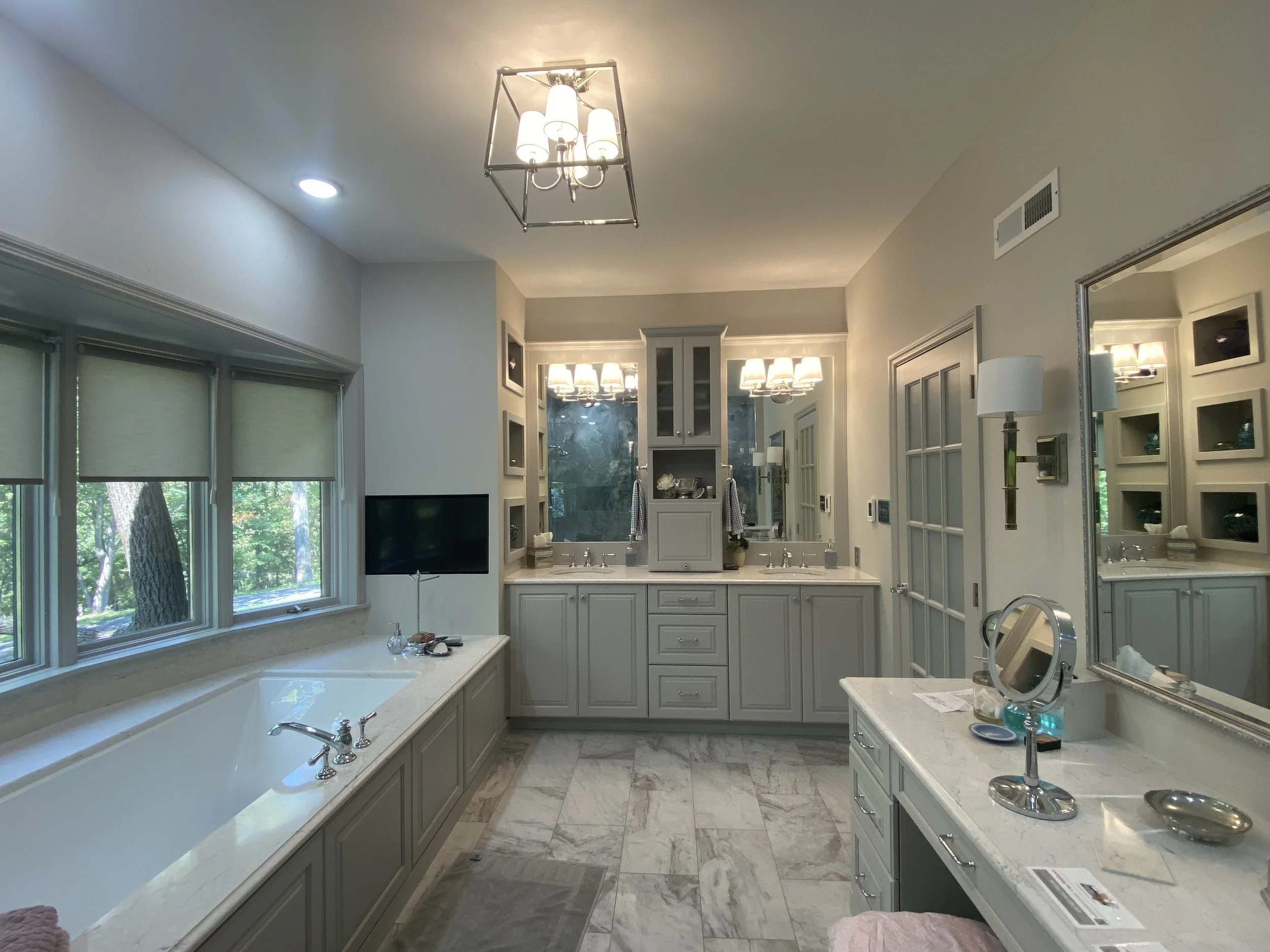 Interior of a bright bathroom with white cabinetry, a bathtub, large windows with blinds, and a marble floor, with a chandelier on the ceiling.