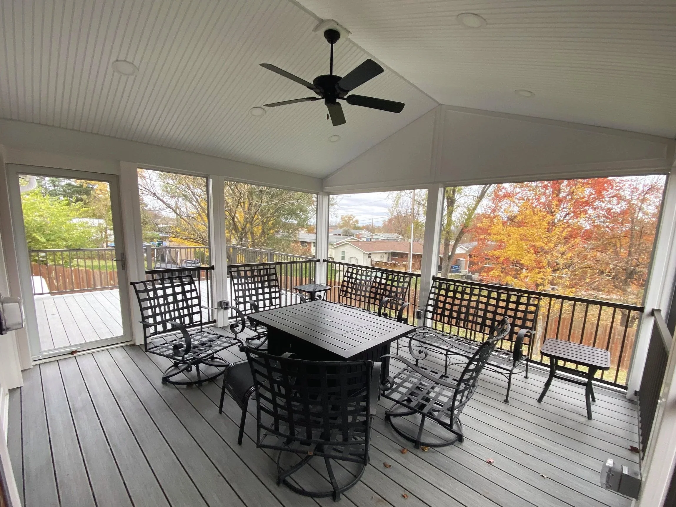 Covered porch or sunroom with ceiling fan, black metal table and chairs, surrounded by windows showing autumn trees and neighboring houses.
