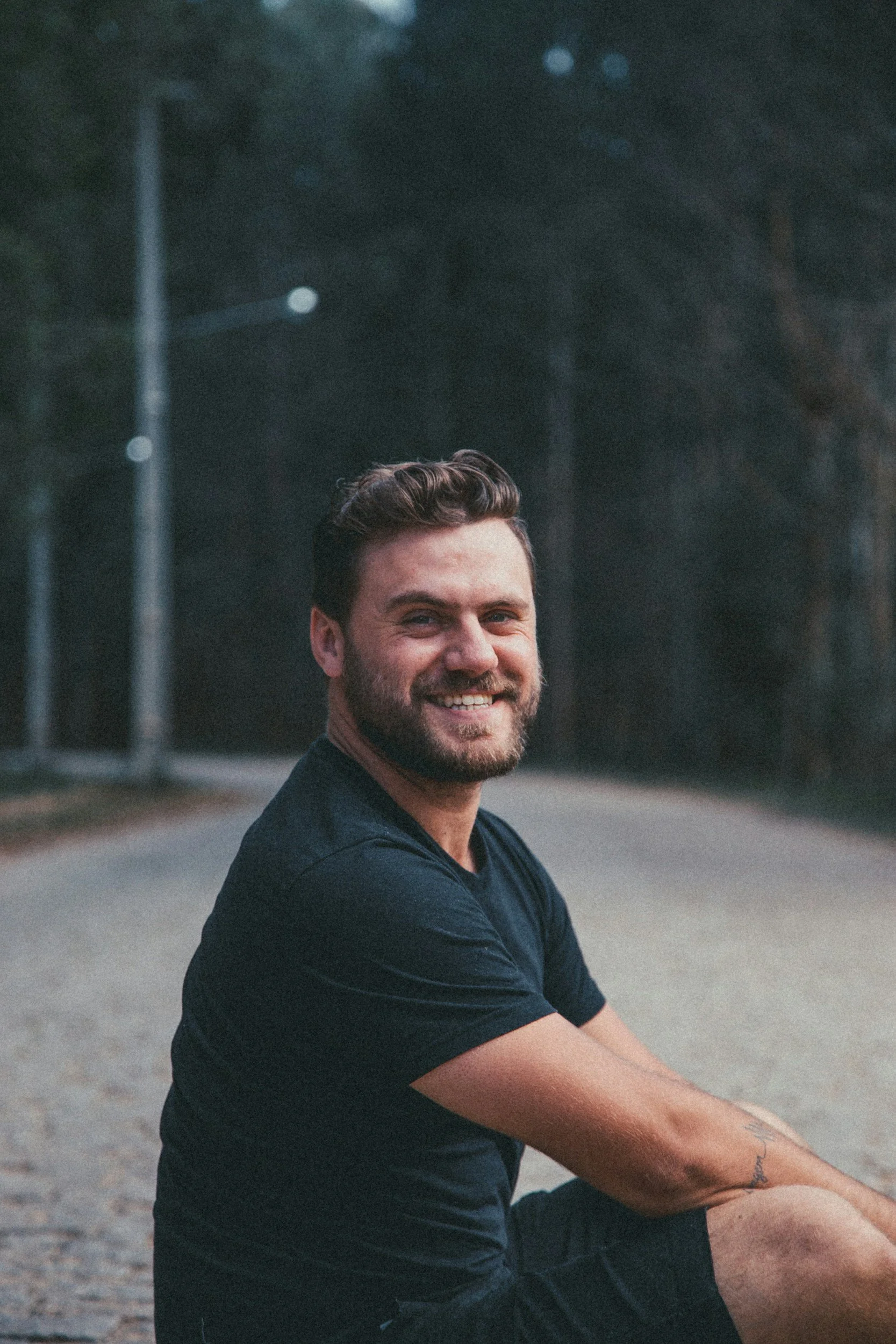 A smiling man with dark hair and a beard sitting outdoors at dusk or evening, wearing a black T-shirt, with a blurred background of trees and streetlights.