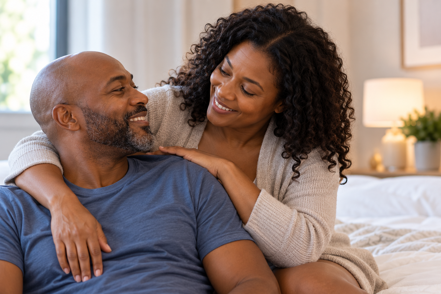 A woman and a man sitting on a bed, smiling and looking at each other affectionately in a cozy bedroom with natural light.