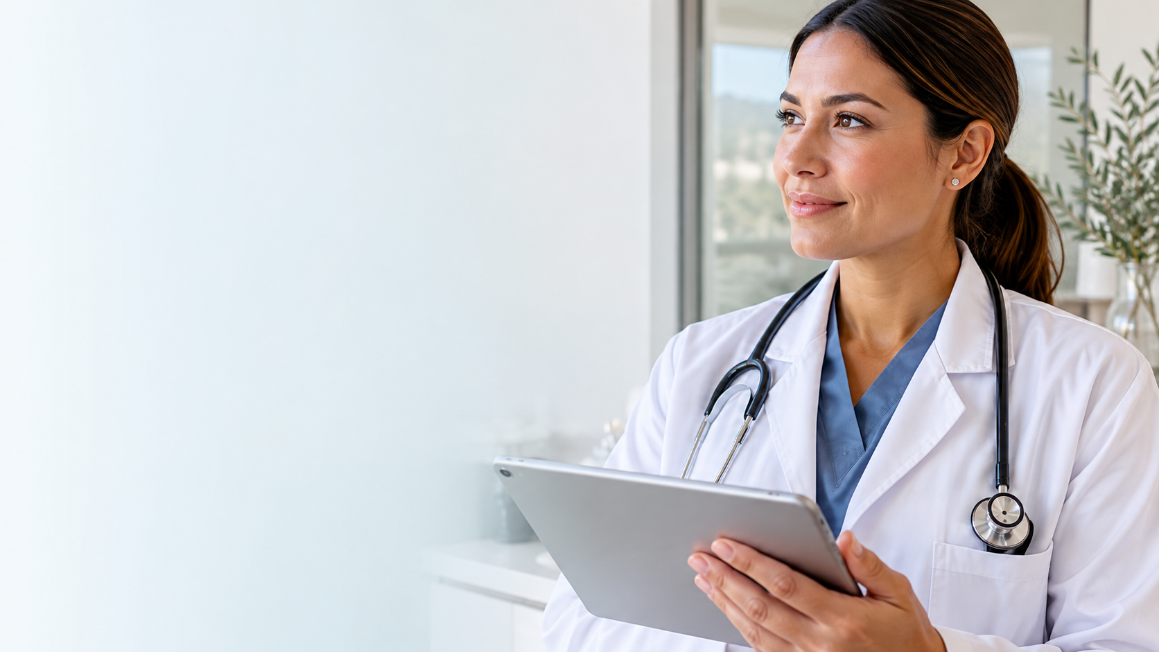 A female doctor with dark hair in a ponytail, wearing a white coat and stethoscope, holding a tablet, smiling, in a bright medical office with large windows and plants.