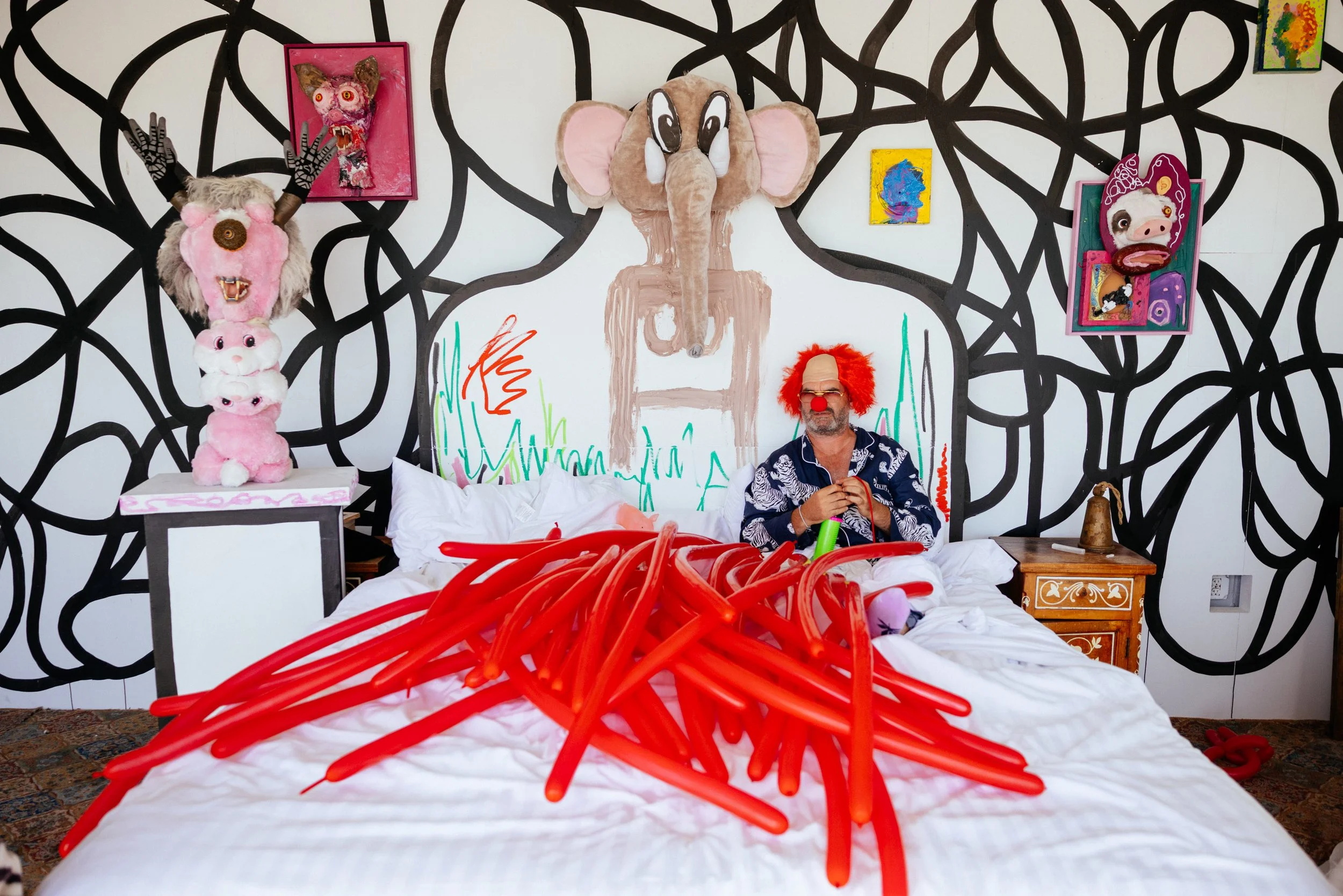 A room with a bed and eclectic artwork. On the wall are three large, colorful, and expressive pieces, including a pink framed monster mask, a yellow and blue abstract portrait, and a pig mask with pink curly hair. The bed has a person sitting at the 