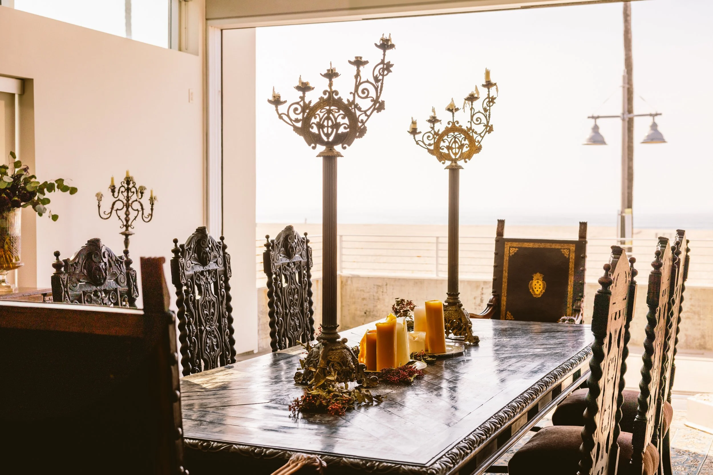 Decorative dining room with a large wooden table, ornate chairs, candles, and candelabras. Large window shows the outside with clear sky and power lines.