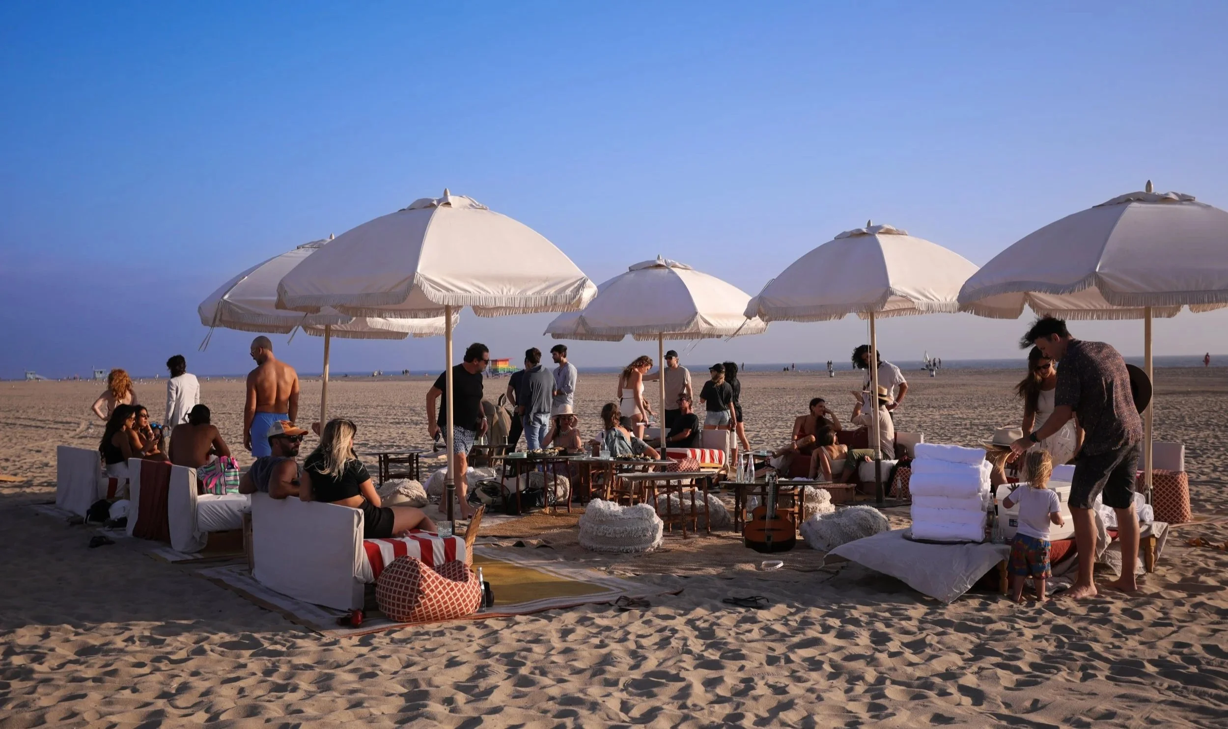 People gathered on the beach under large white umbrellas, sitting on outdoor furniture, socializing and enjoying the sunset.
