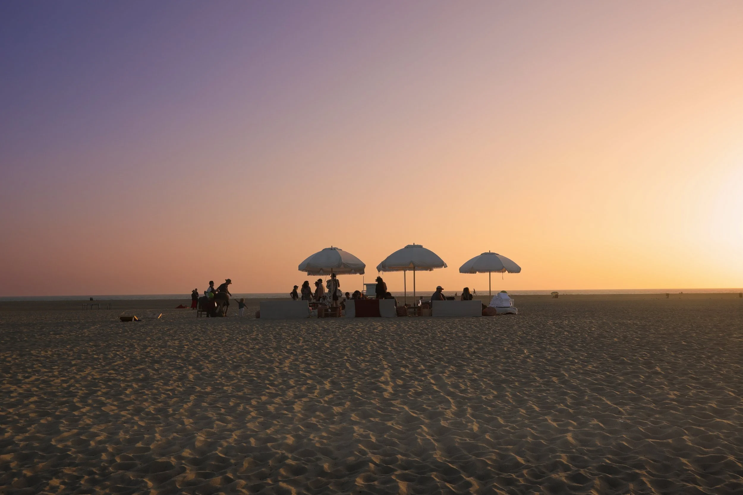 Beach scene during sunset with groups of people, umbrellas, and lounge chairs on the sandy shore.
