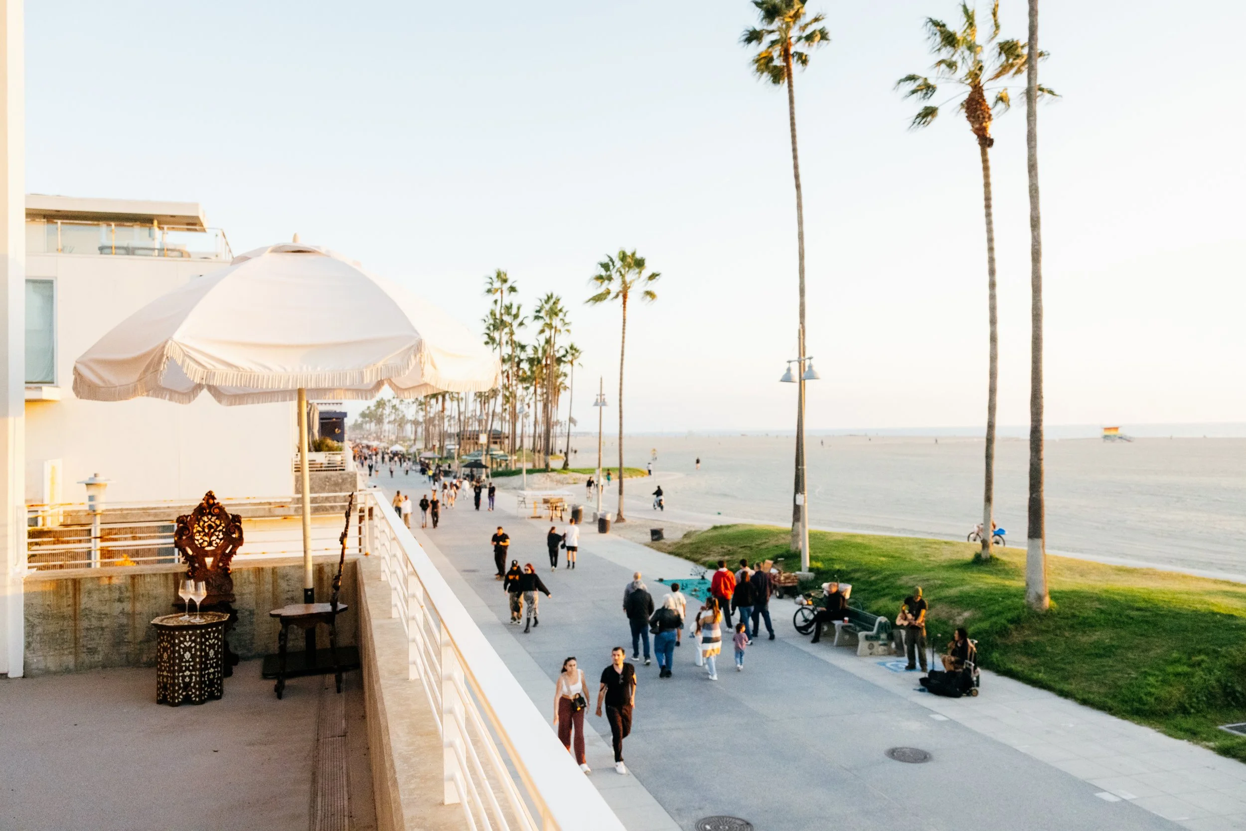 People walking along a boardwalk by the beach on a sunny day in Los Angeles, California, with palm trees and a lifeguard tower in the distance.