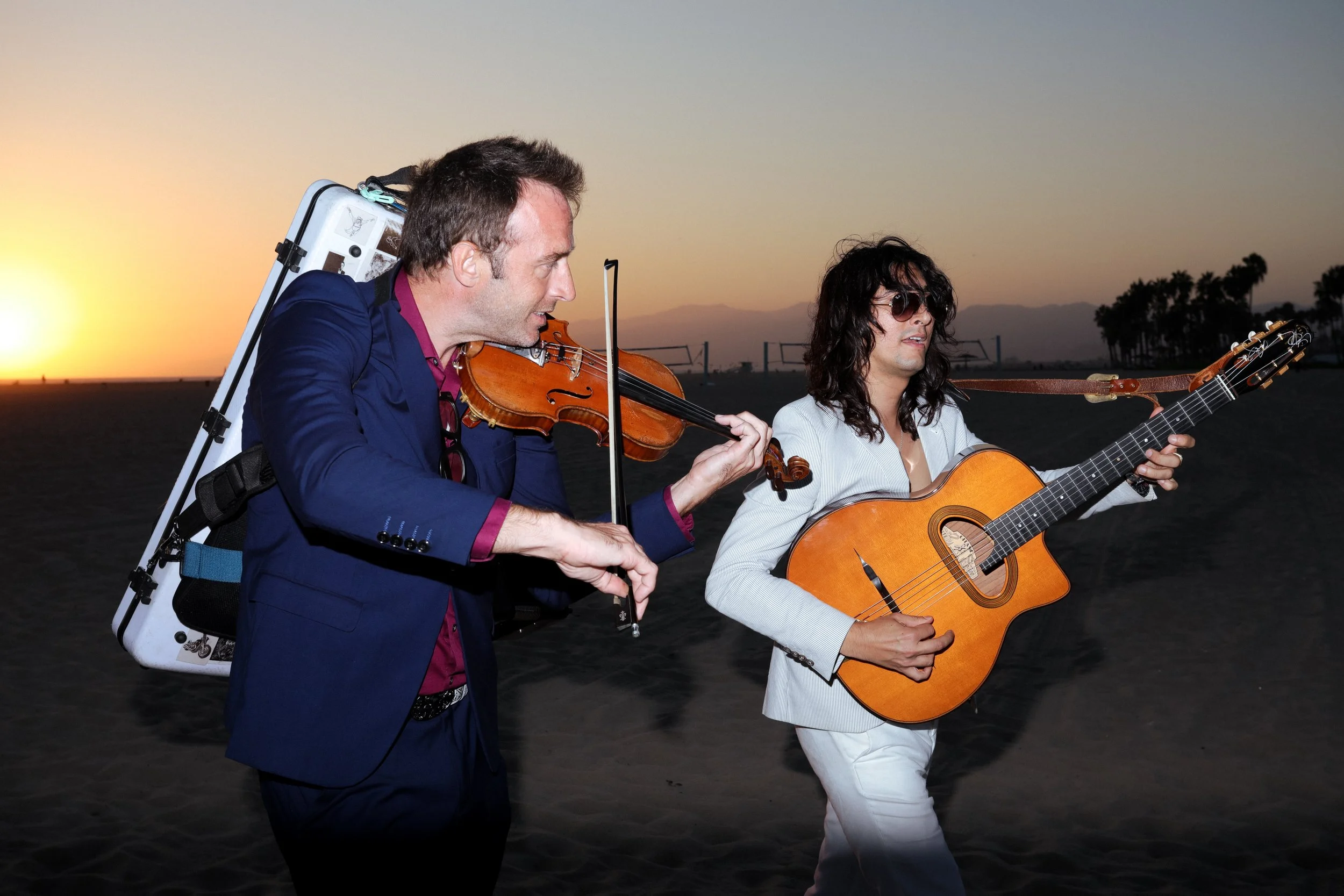 Two musicians performing on a beach at sunset, one playing the violin and the other playing the guitar.